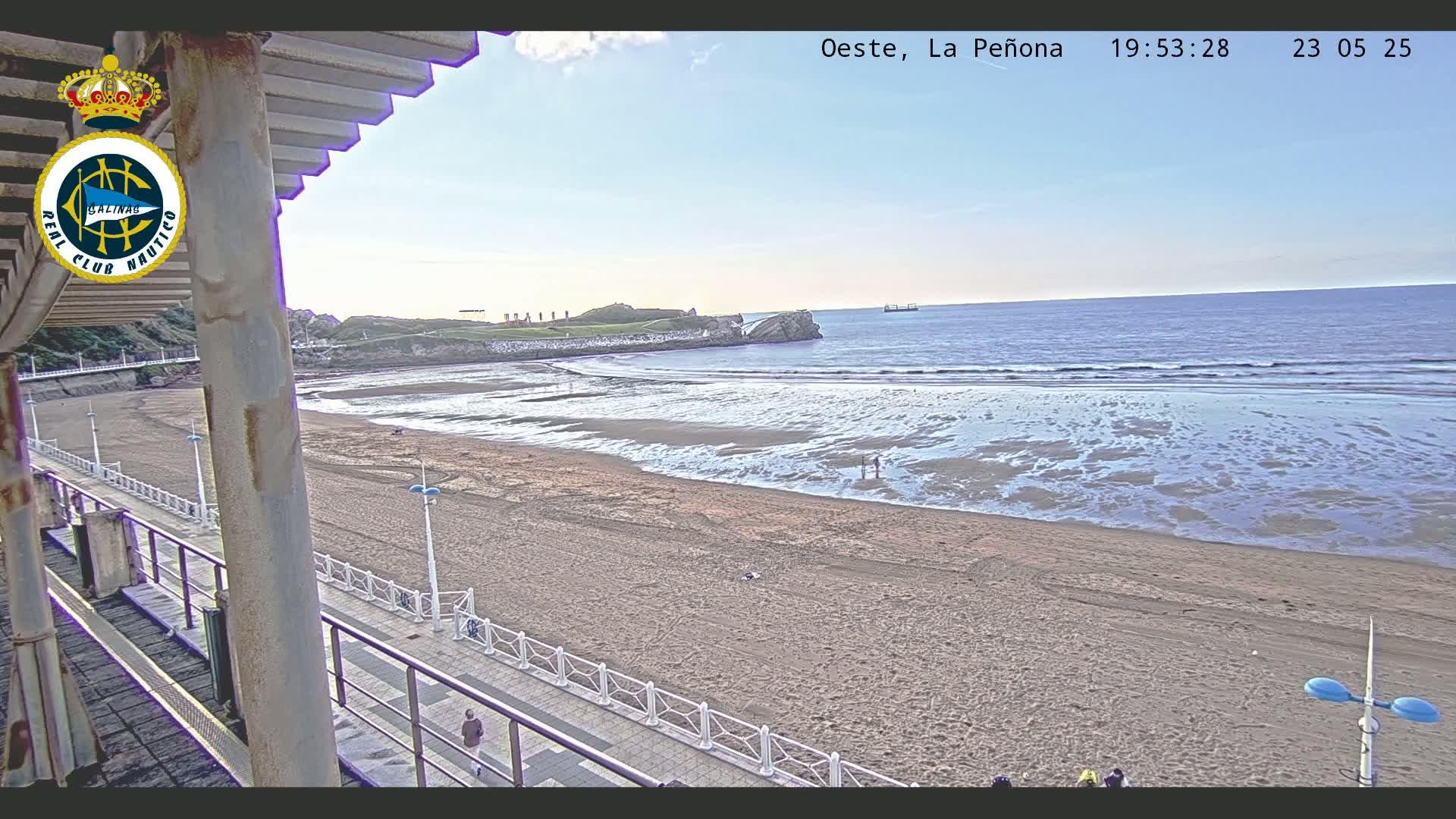 A sandy beach stretches out to a calm ocean under a clear, light-blue sky, viewed from an elevated vantage point.