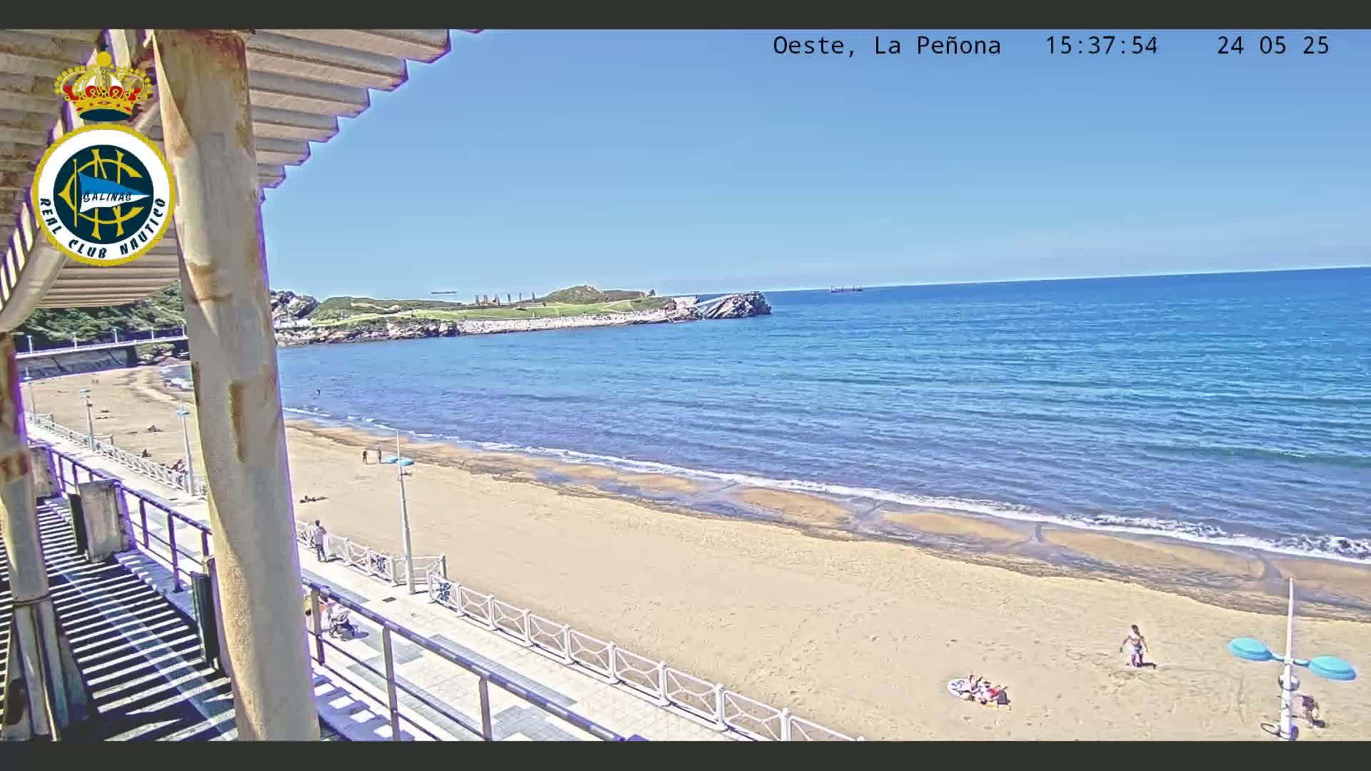 A sandy beach with a calm ocean under a clear blue sky is seen from a high vantage point, near a walkway and some buildings.