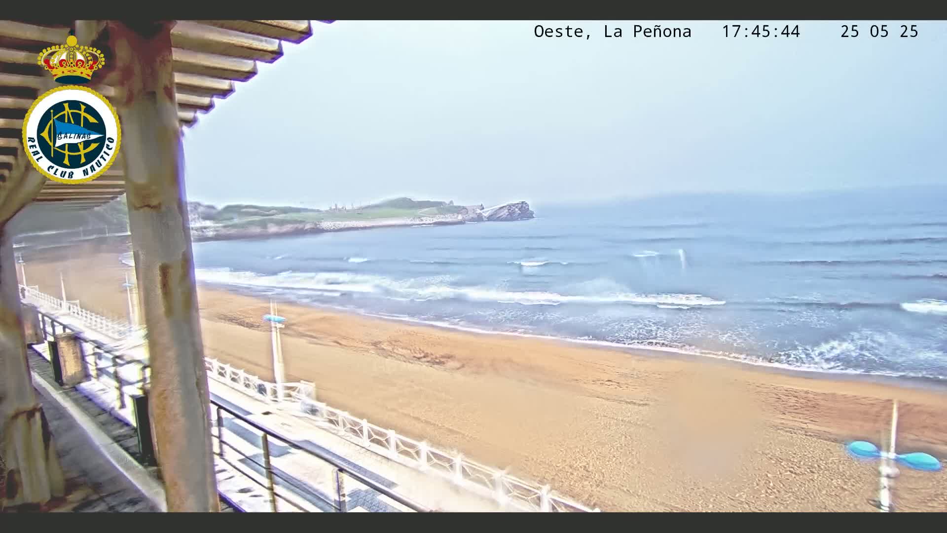 A sandy beach with moderate waves under a hazy sky is viewed from a covered balcony.
