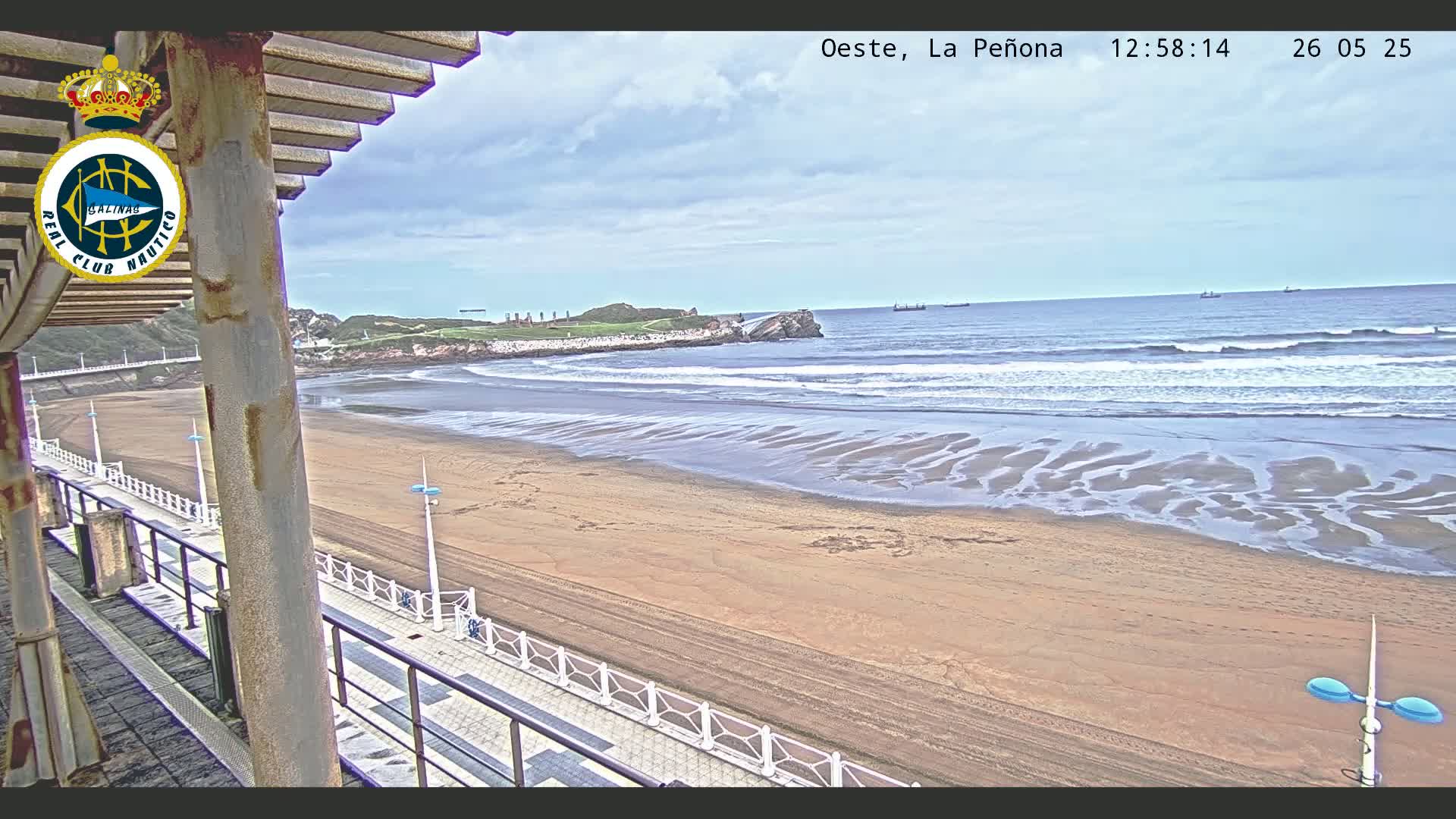 A sandy beach with gentle waves under a partly cloudy sky is viewed from an elevated position, showing a boardwalk and a portion of a building.