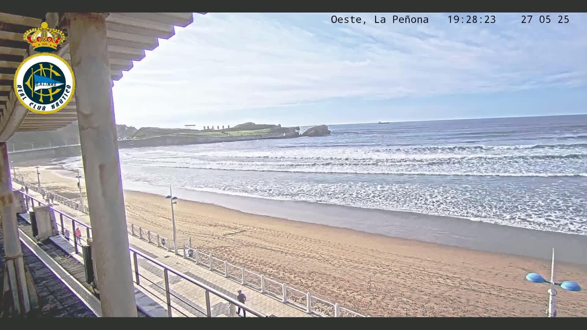 A sandy beach with gentle waves rolling onto the shore on a partly sunny day, viewed from a seaside structure.