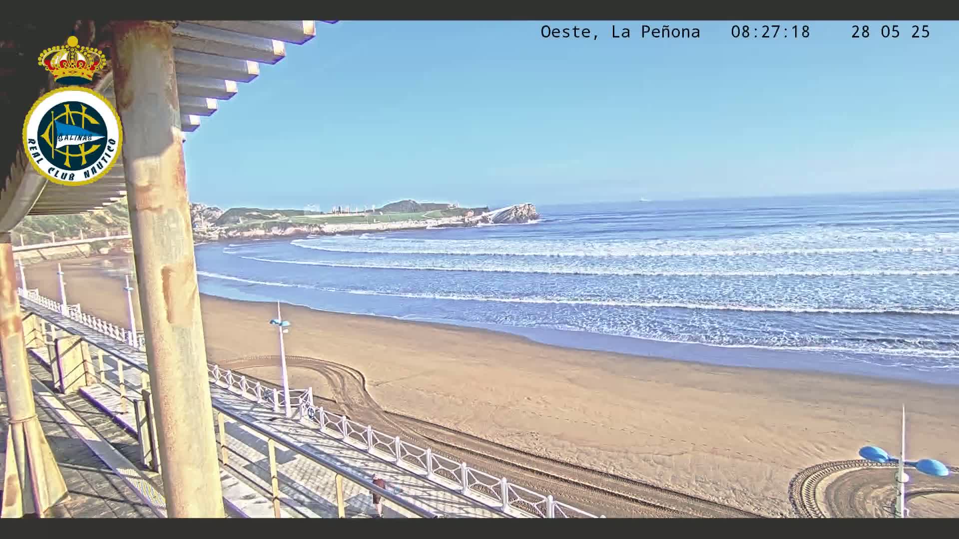 A sandy beach with gentle waves under a clear blue sky is viewed from an elevated vantage point, showing a promenade and part of a building.
