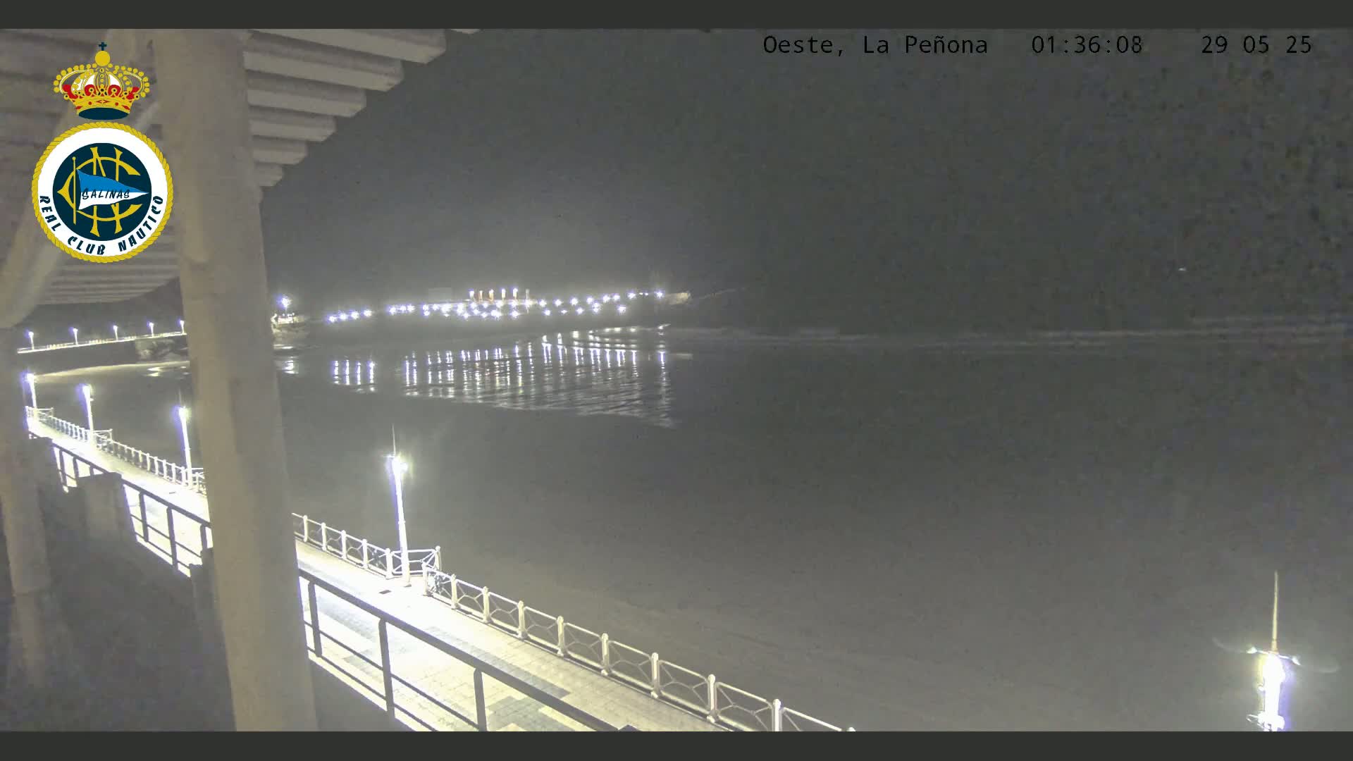 A nighttime view from a pier shows a calm, dark ocean reflecting the lights of a beachfront at low tide.