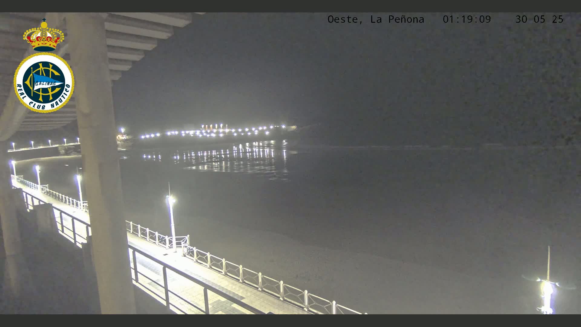 A nighttime view from a pier shows a calm body of water reflecting the lights of a distant shoreline.