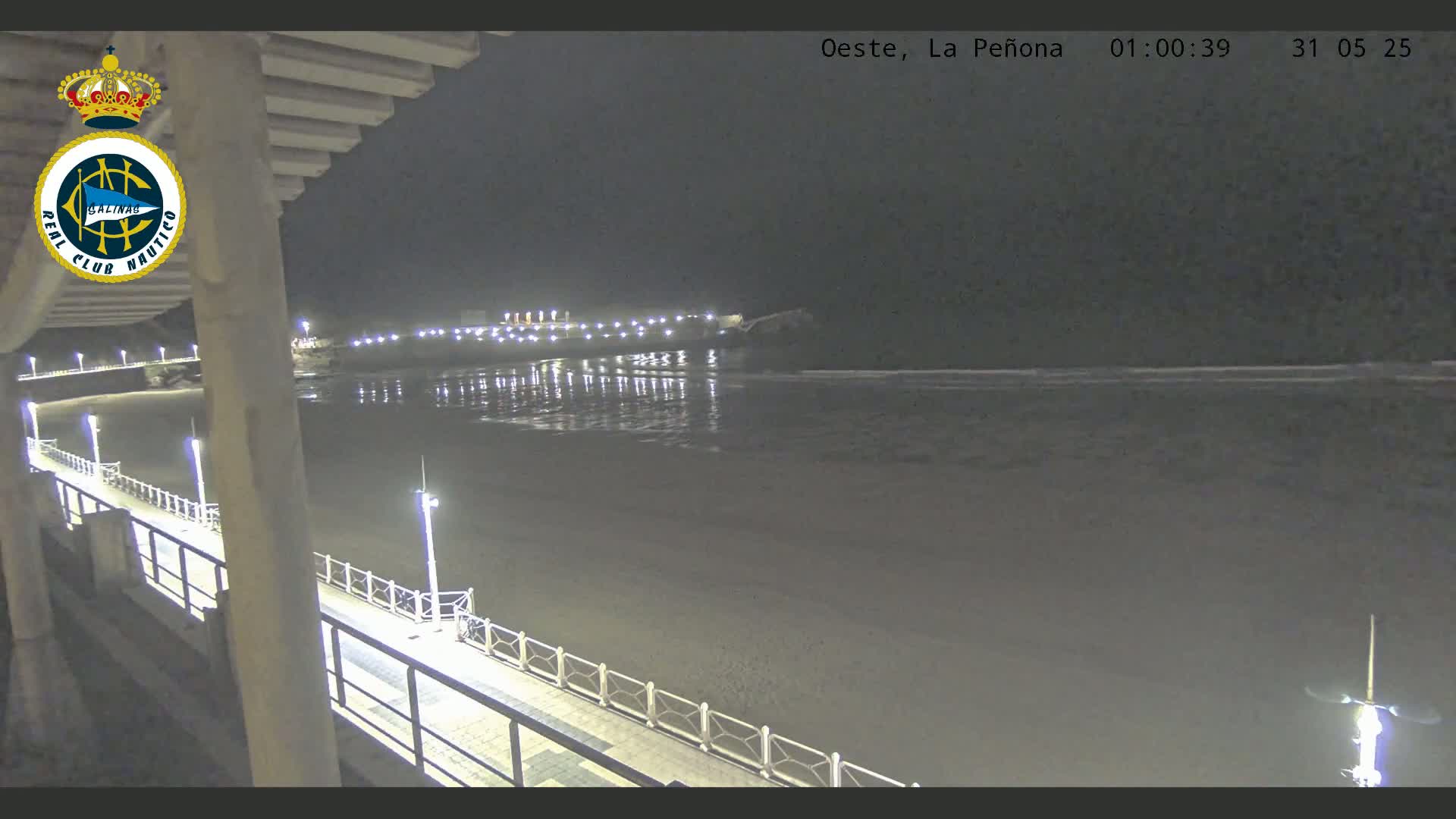 A nighttime view from a pier shows a calm, dark ocean reflecting lights from a shoreline at a beach.