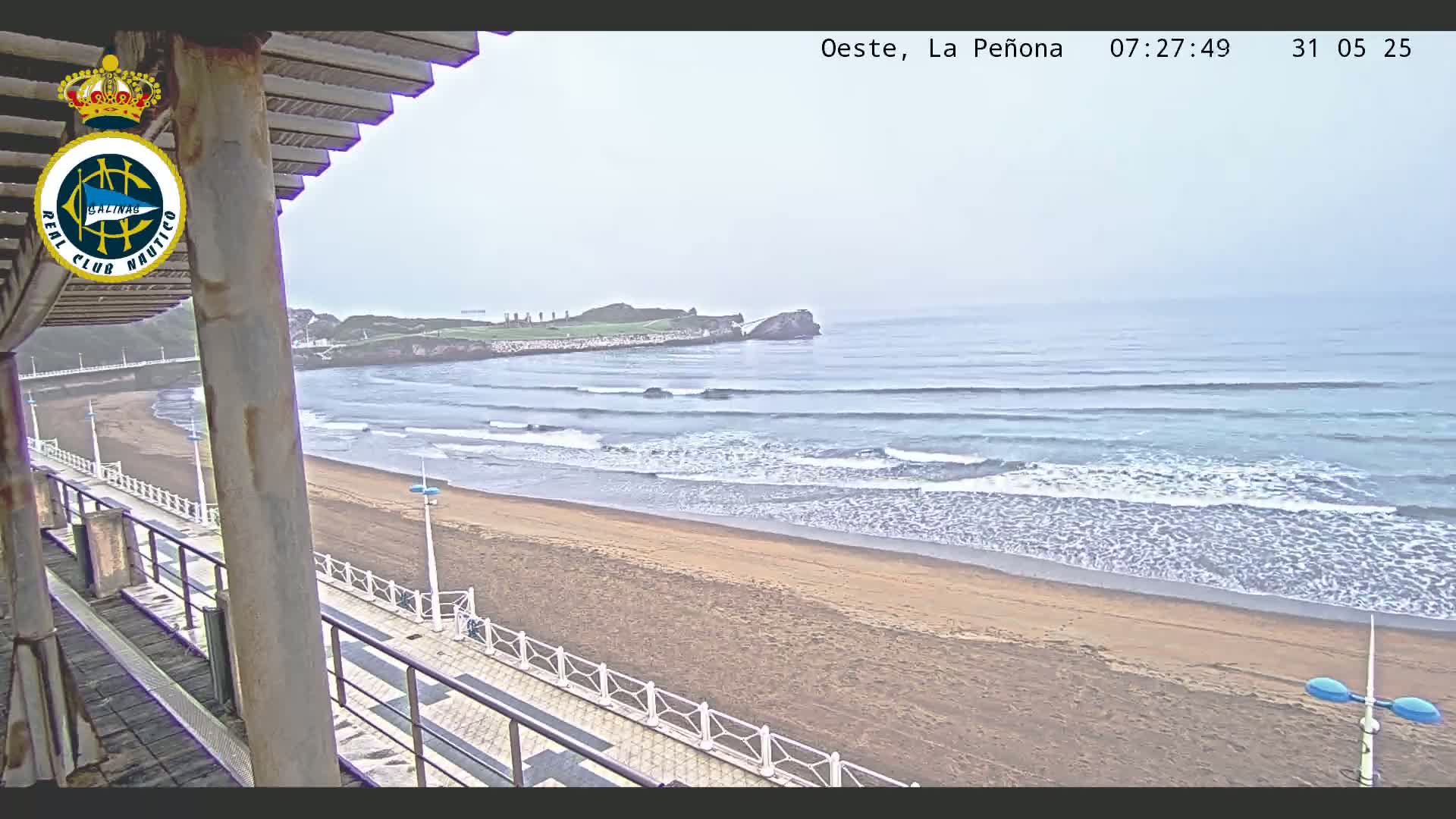 A sandy beach with gentle waves under an overcast sky is viewed from an elevated position, overlooking a paved walkway and a partial view of a building.