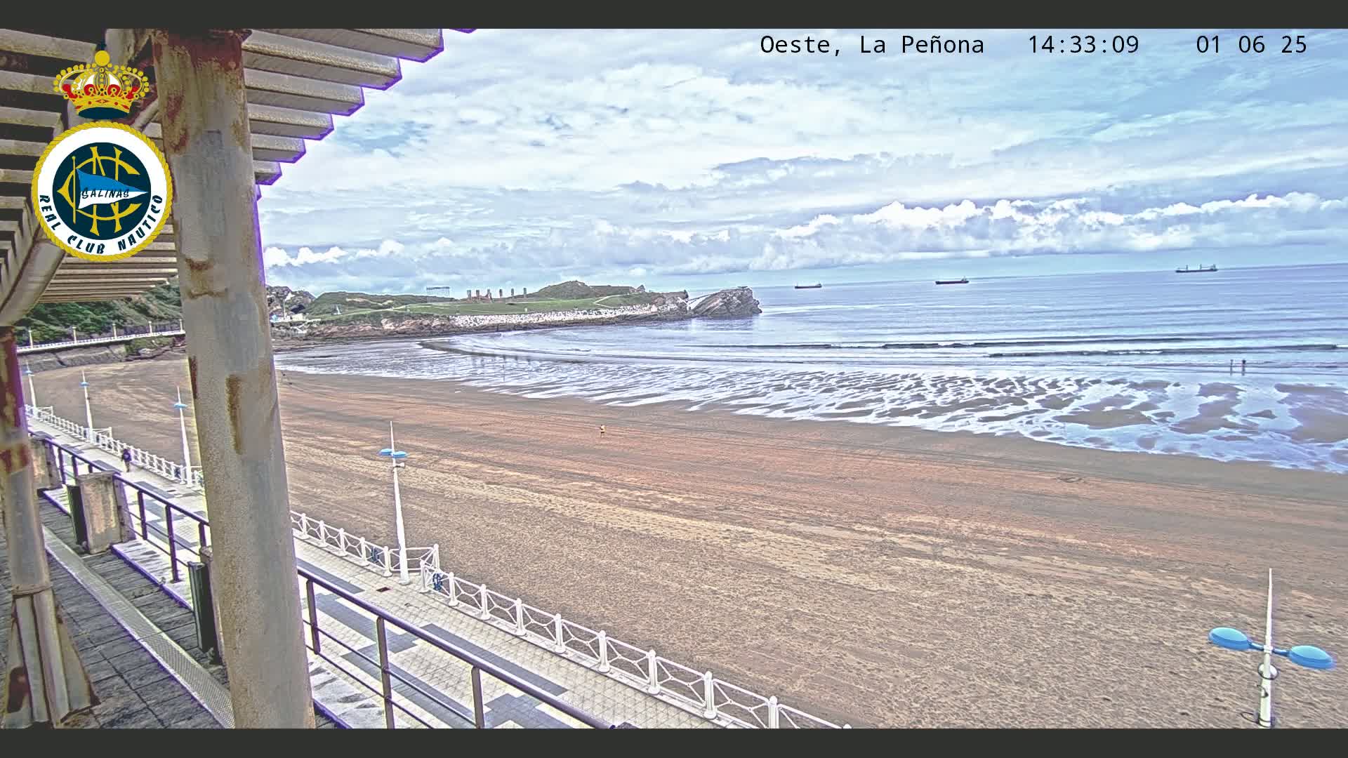 A sandy beach with gentle waves under a partly cloudy sky is viewed from an elevated position, showing a boardwalk and distant ships at sea.