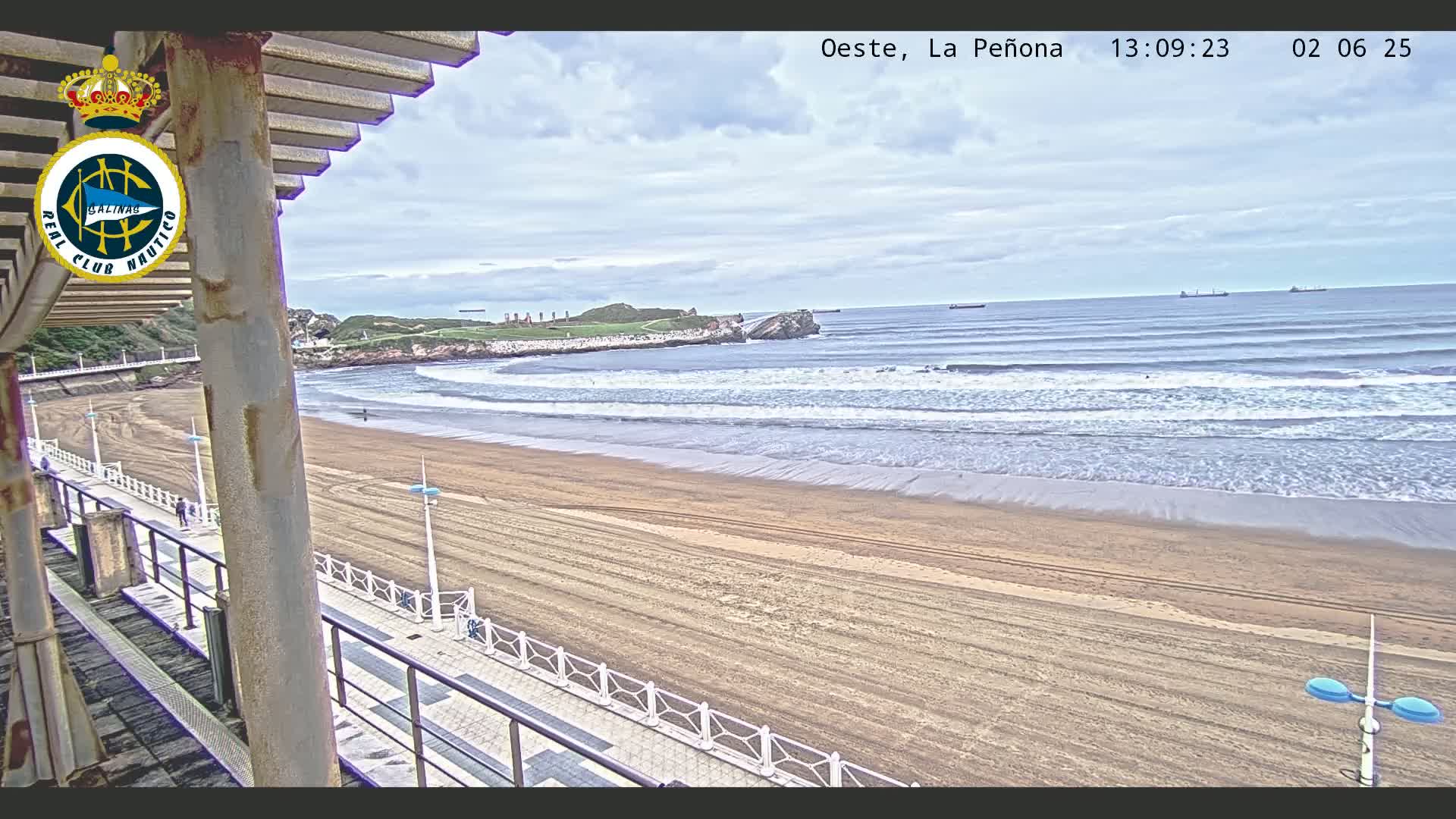 A mostly cloudy day shows a wide sandy beach with gentle waves rolling onto the shore, viewed from an elevated position overlooking a boardwalk.