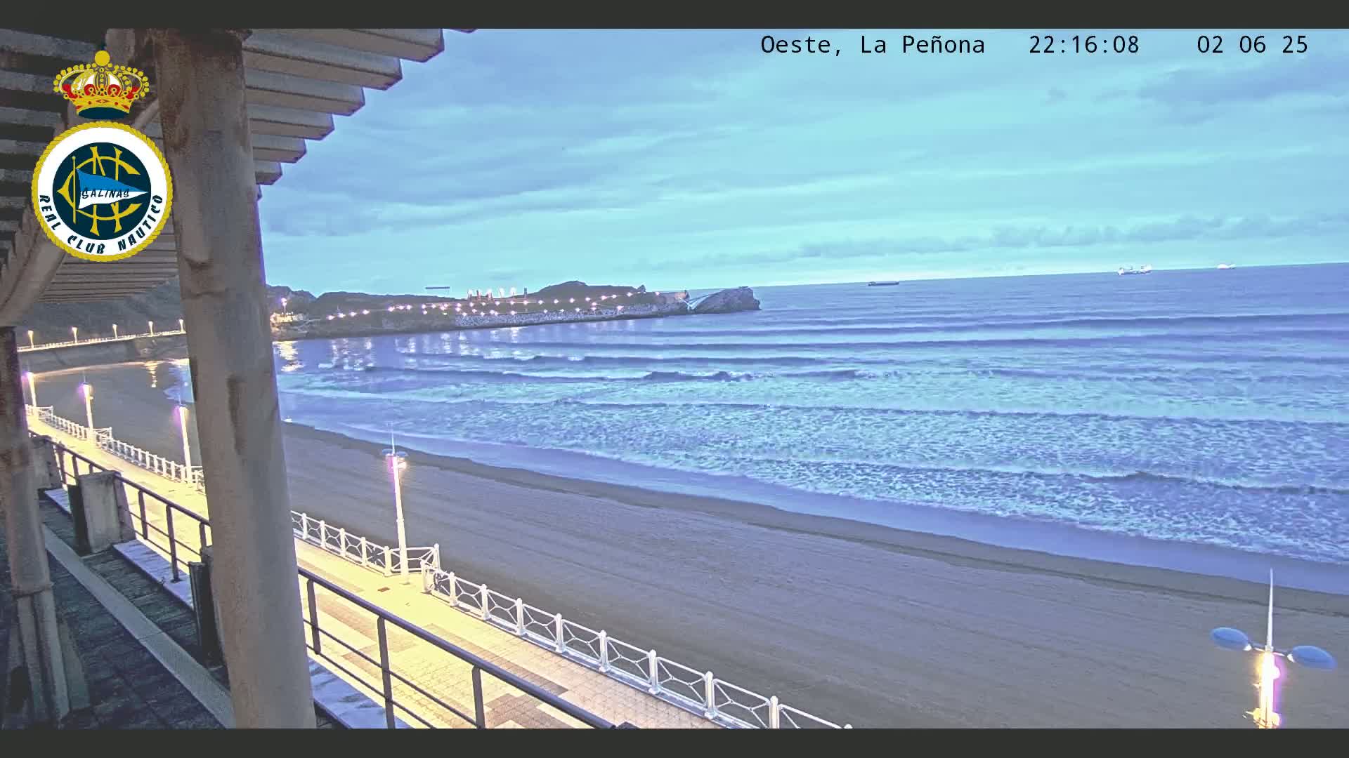 A mostly clear twilight sky overlooks a sandy beach with gentle waves rolling in, lit by lamps along a boardwalk.