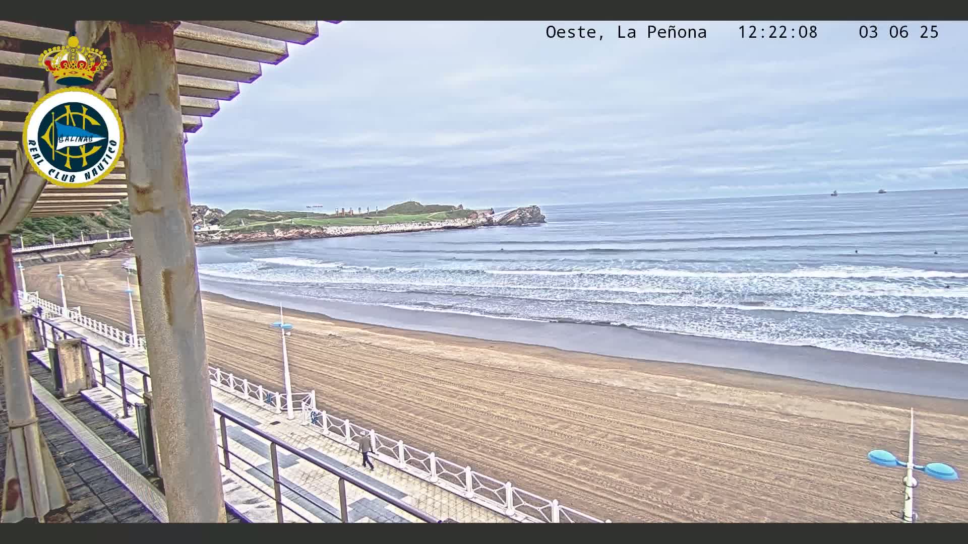 A sandy beach with gentle waves under an overcast sky is viewed from a partially covered elevated walkway.