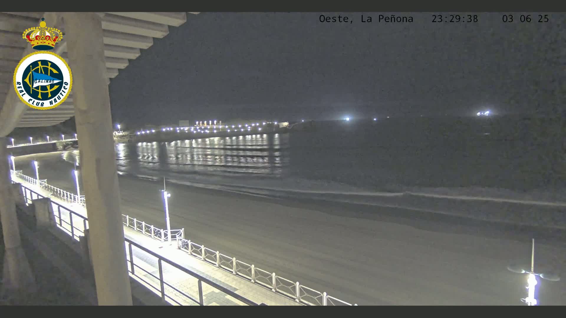 A nighttime view from a balcony overlooking a dark sandy beach with gently lapping waves, a lit pier in the distance, and a boardwalk illuminated by streetlights.