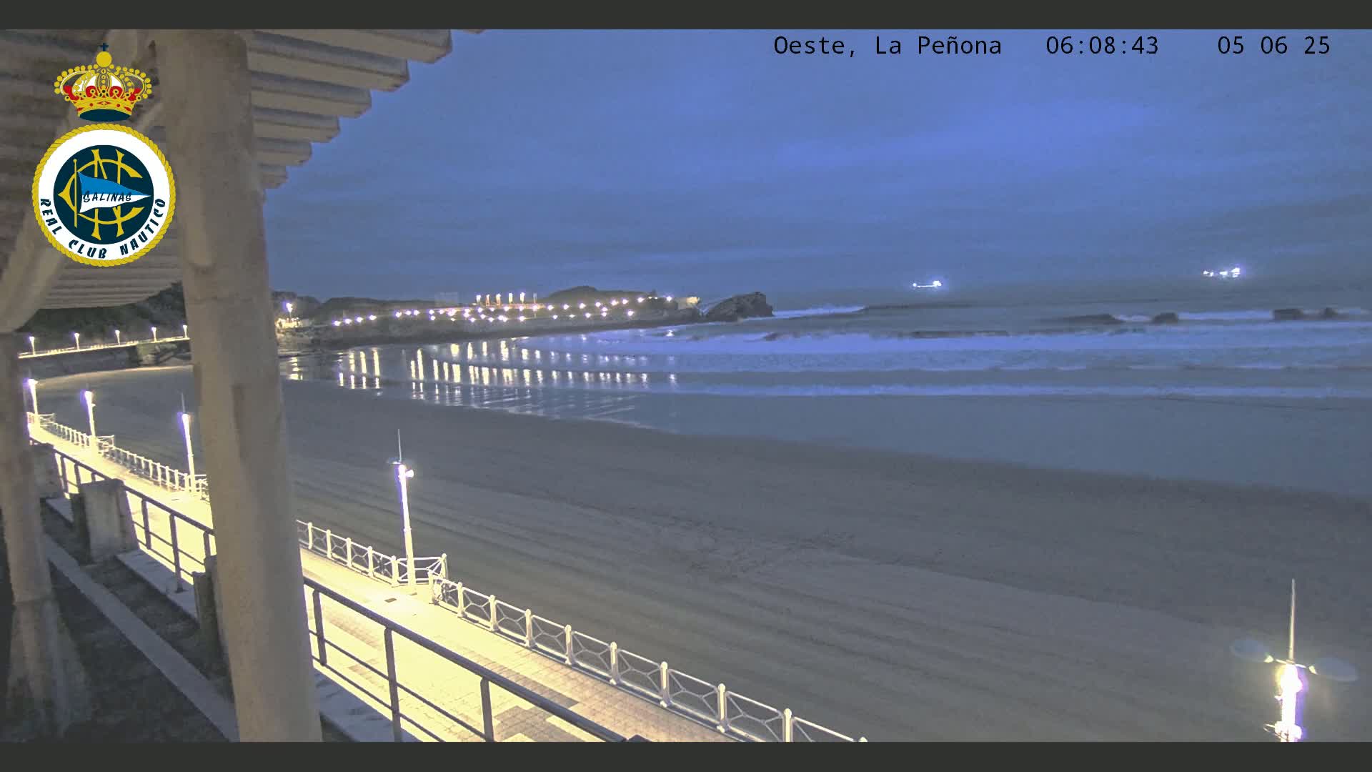 A dark, twilight view of a beach at night with calm waves lapping the shore, illuminated by lights along a boardwalk.
