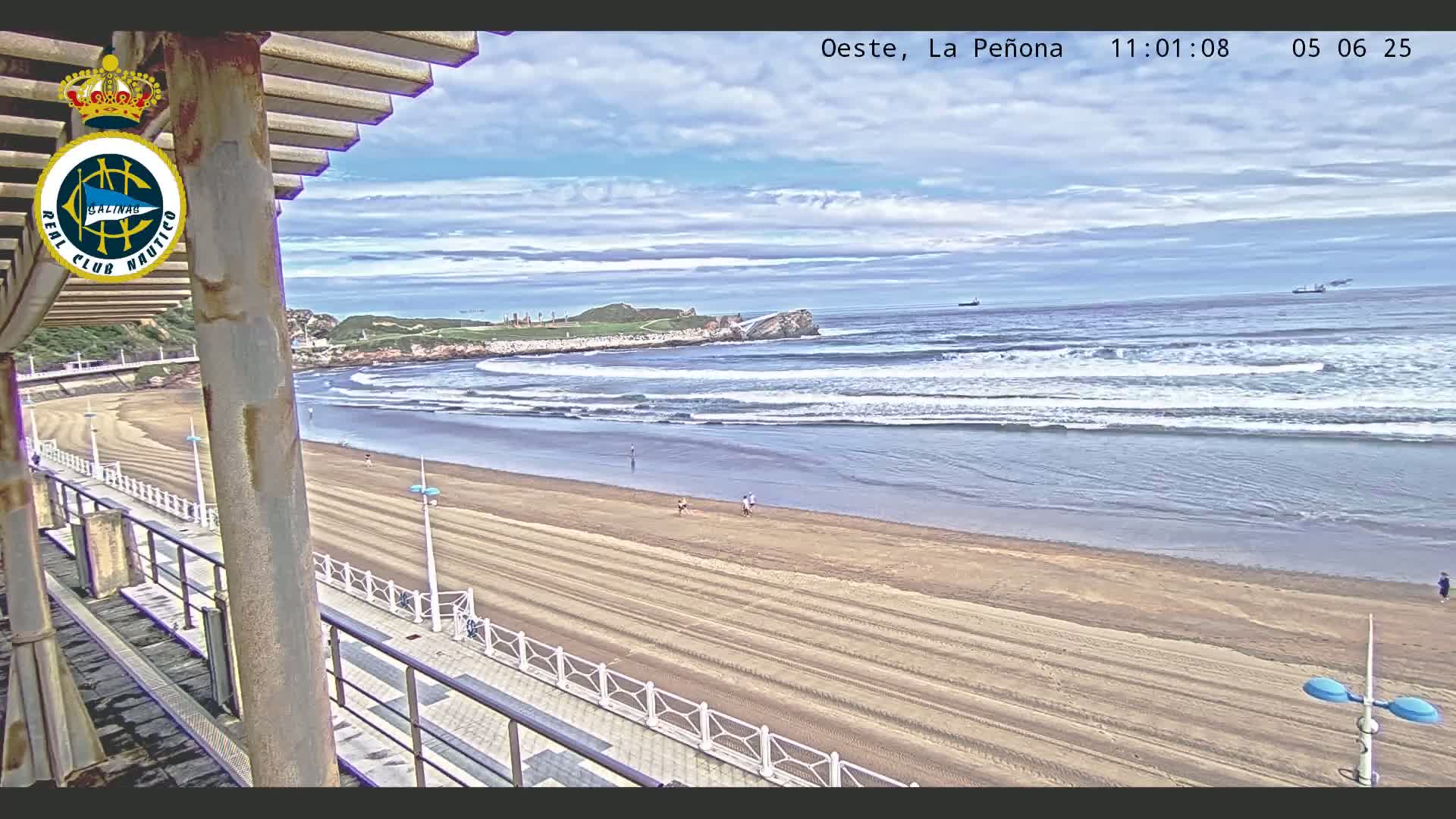 A sandy beach with a few people on it is seen from an elevated viewpoint, with waves breaking in the ocean under partly cloudy skies.