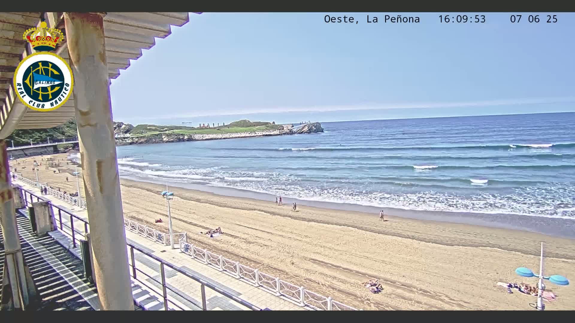 A sandy beach with a few people on it, gentle waves rolling onto the shore under a clear blue sky, is viewed from an elevated position.
