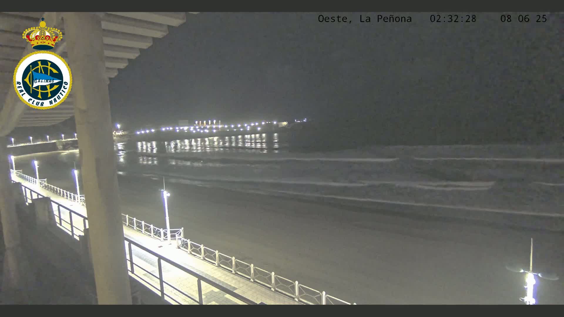 A nighttime view from a pier shows a dark beach with gentle waves lapping the shore under a clear sky, and a line of lights visible in the distance.