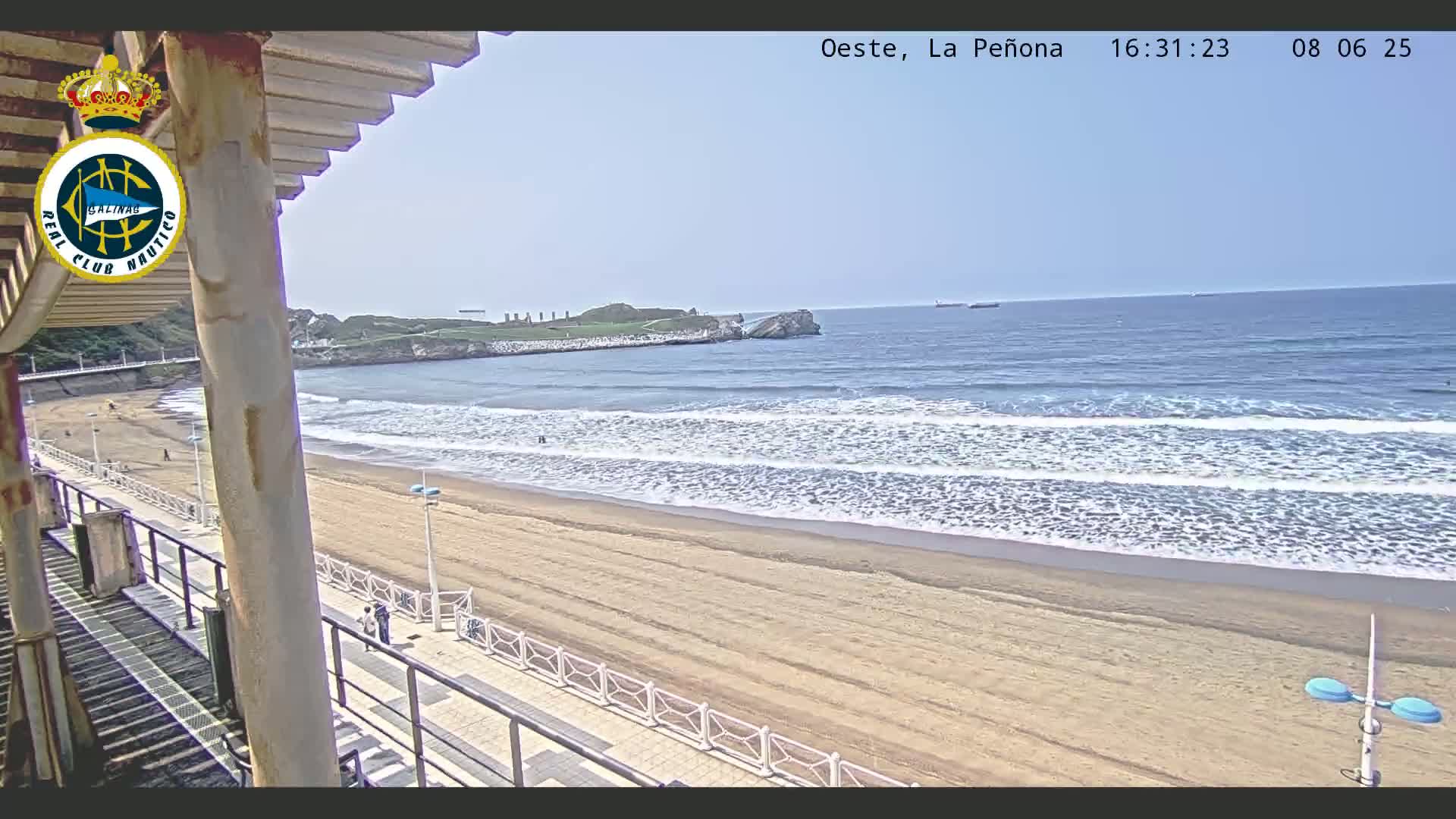 A sandy beach with gentle waves laps the shore under a clear sky, viewed from a structure overlooking the ocean.