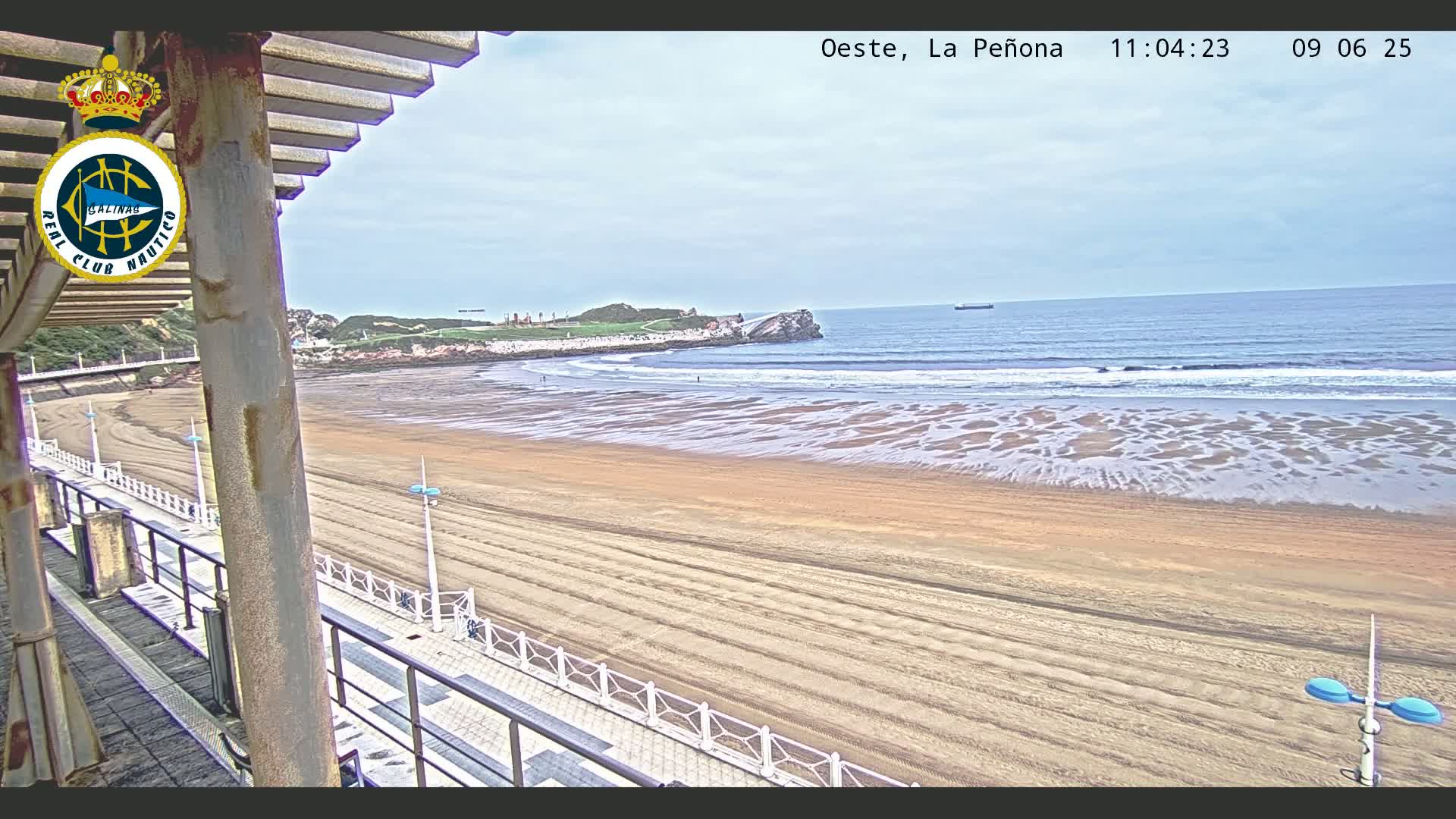 A sandy beach stretches out to a calm ocean under a mostly cloudy sky, viewed from an elevated vantage point with a boardwalk and railing in the foreground.