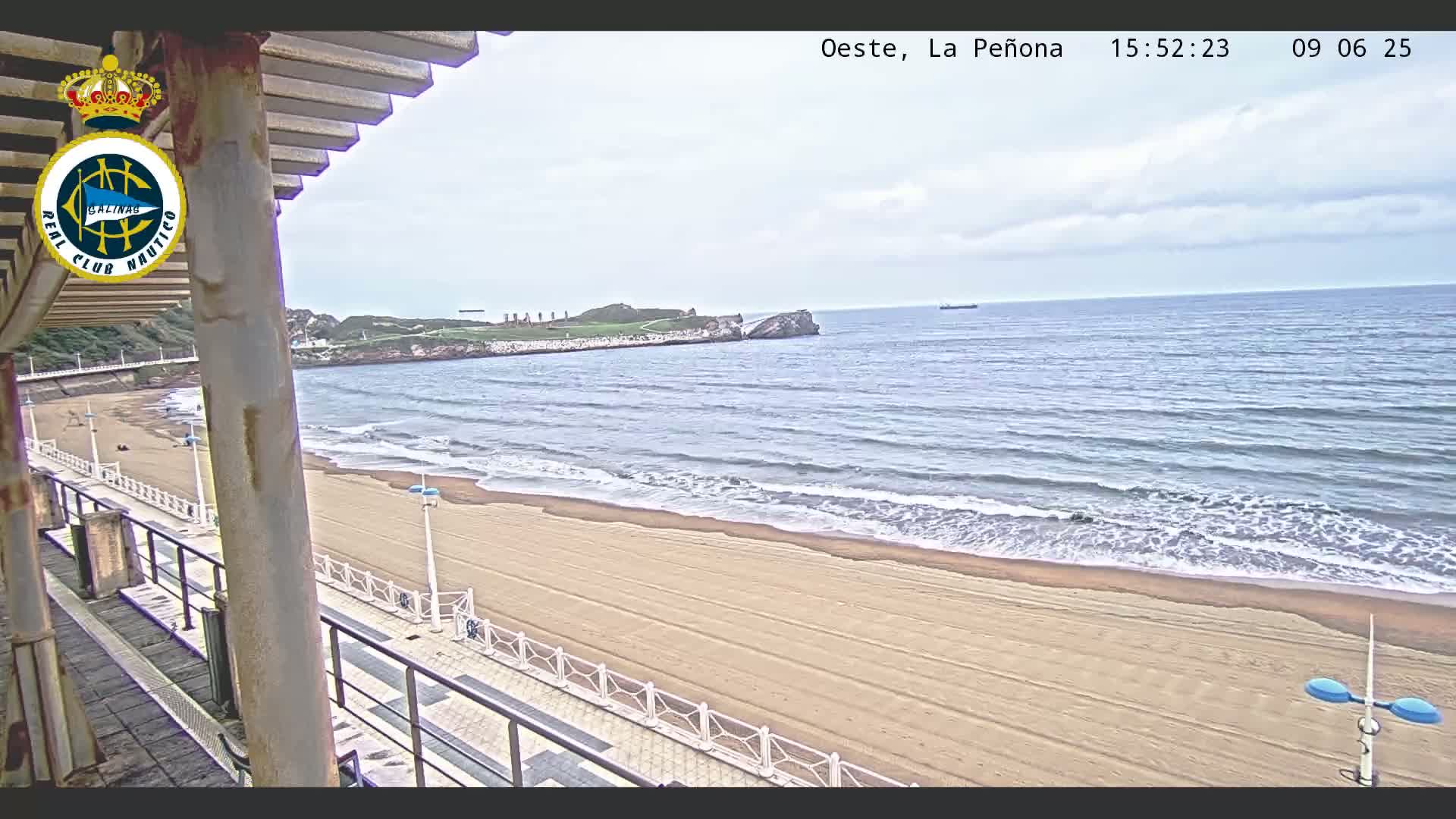 A sandy beach with gentle waves under a cloudy sky is seen from an elevated viewpoint, showing a promenade and a rocky outcropping in the distance.