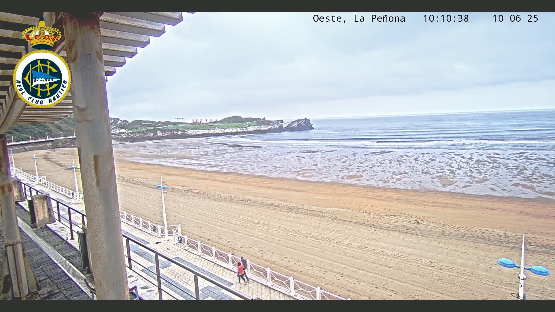 A wide sandy beach stretches out to a calm ocean under an overcast sky, with two people walking along a paved walkway near the water's edge.