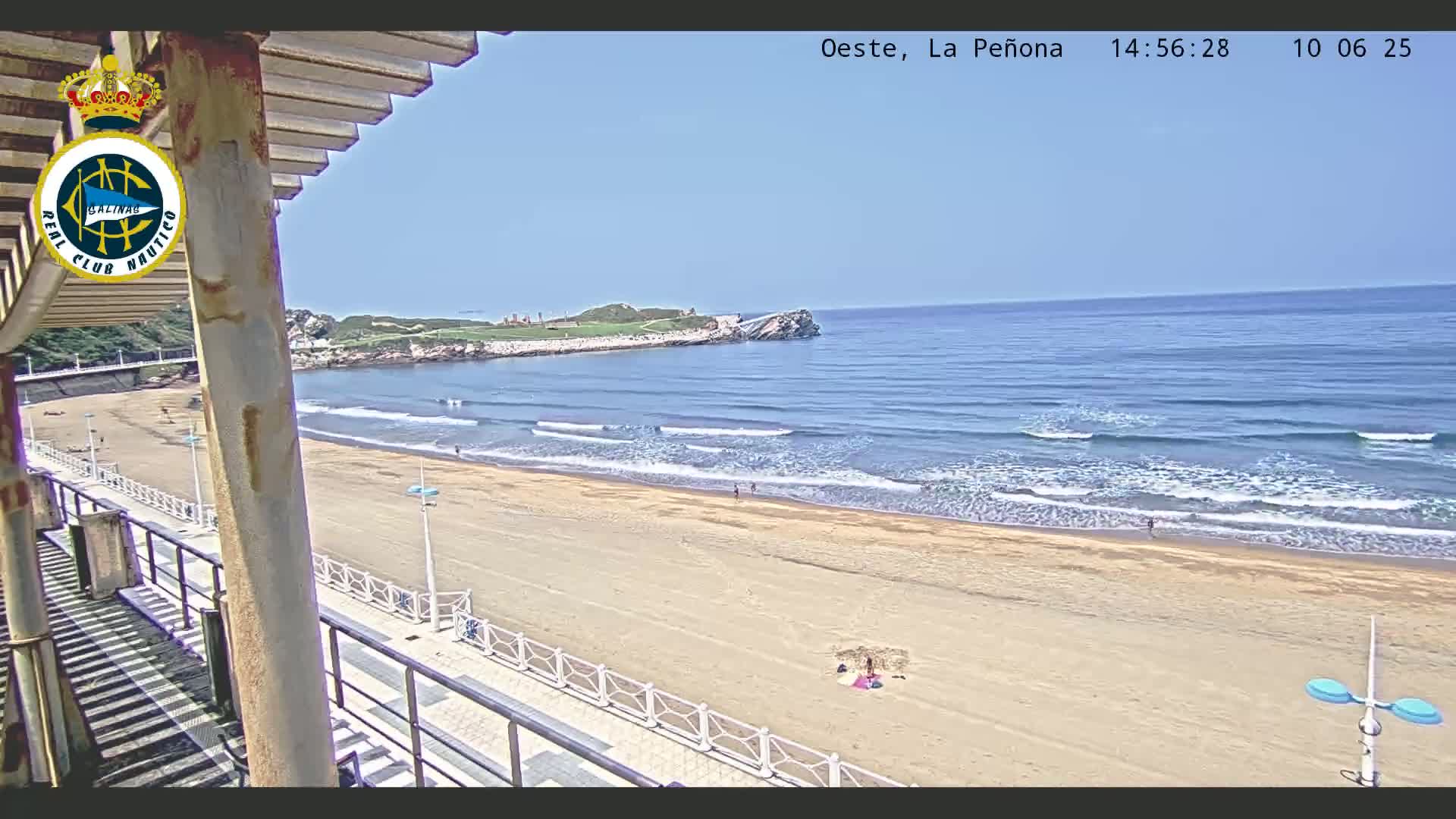 A sandy beach with gentle waves under a clear blue sky is viewed from an elevated position, showing a boardwalk and a small number of people on the beach.