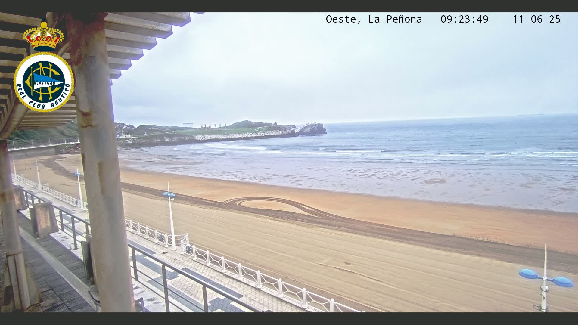 A sandy beach with small waves under an overcast sky is viewed from a balcony overlooking a promenade.