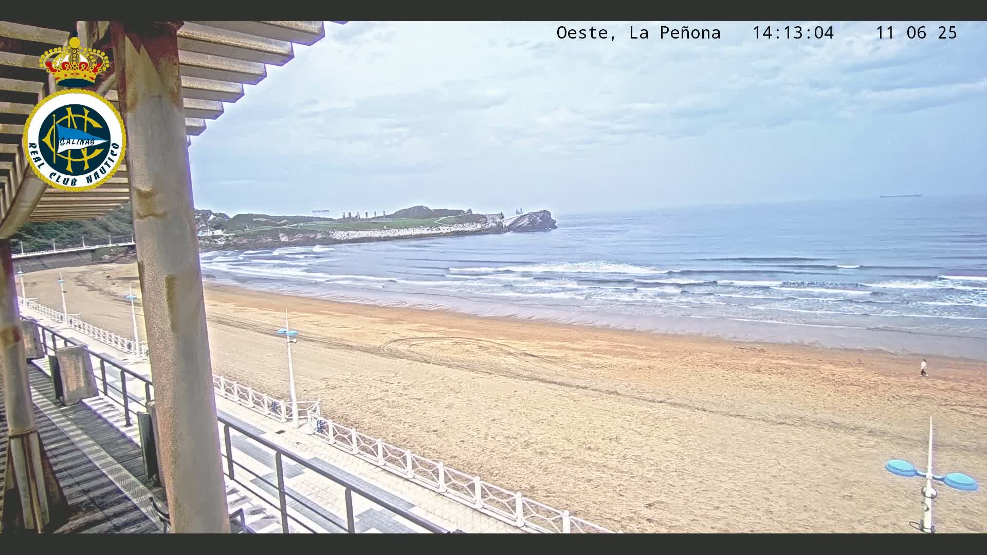A sandy beach with gentle waves under a partly cloudy sky is viewed from an elevated position, showing a boardwalk and a rocky outcrop in the distance.