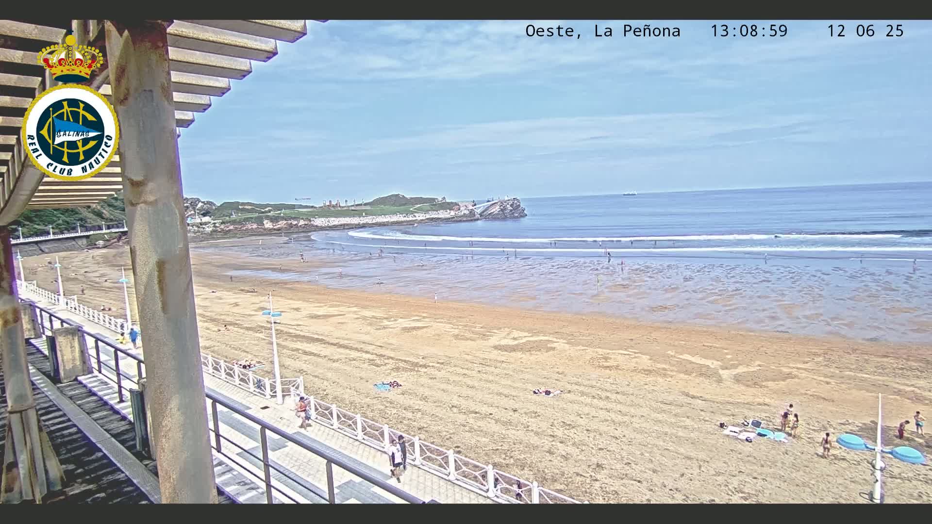A sandy beach with scattered people stretches to a calm ocean under a mostly clear sky, viewed from an elevated perspective near a boardwalk.