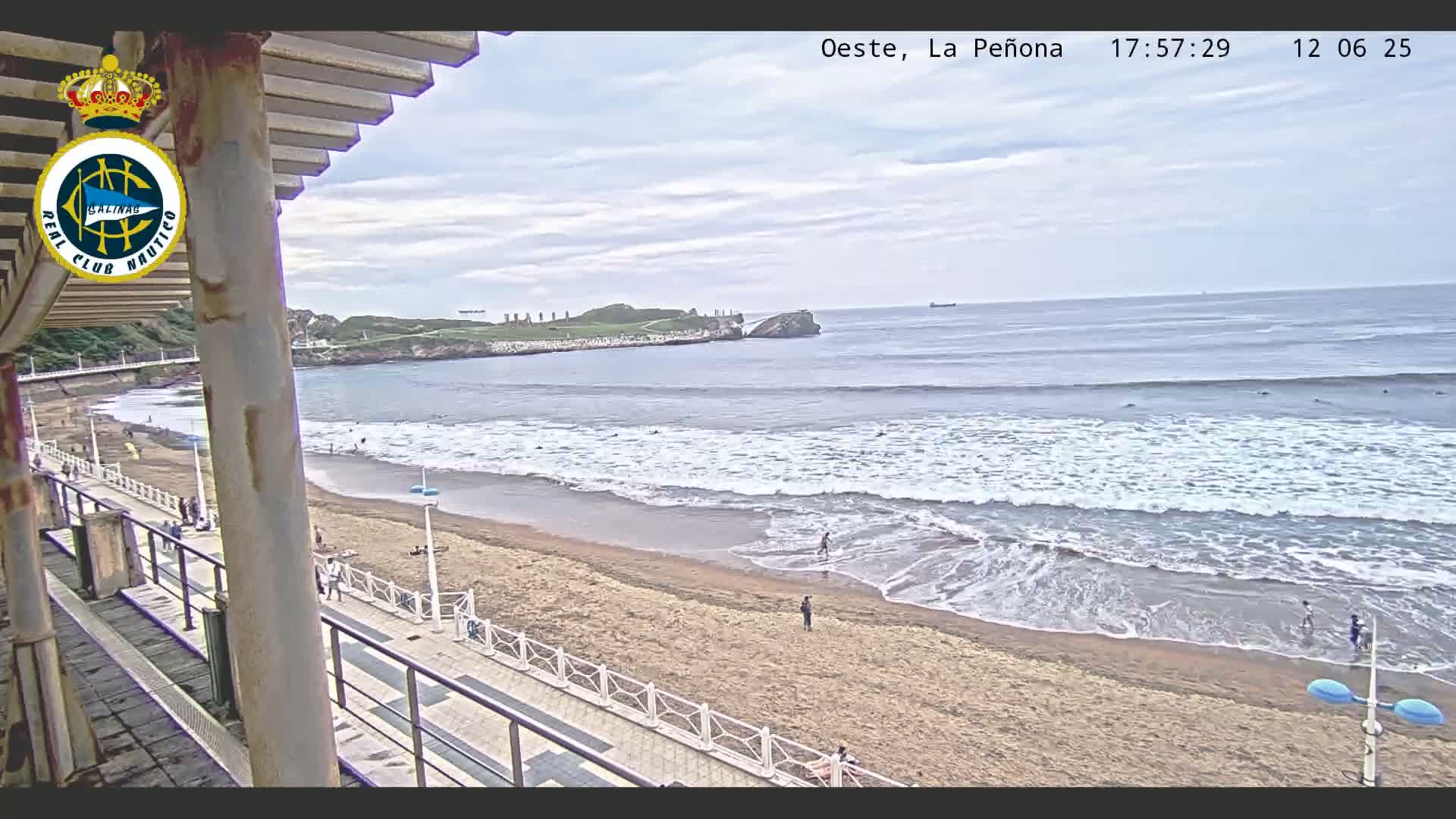 A sandy beach with gentle waves and several people are visible from a partially covered balcony overlooking the ocean on an overcast day.