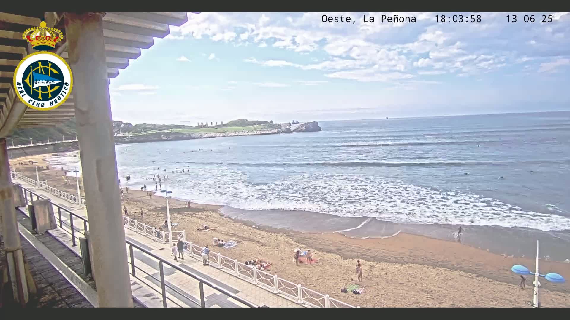 A sandy beach with people sunbathing and swimming is seen from an elevated vantage point, with gentle waves rolling onto the shore under a partly cloudy sky.
