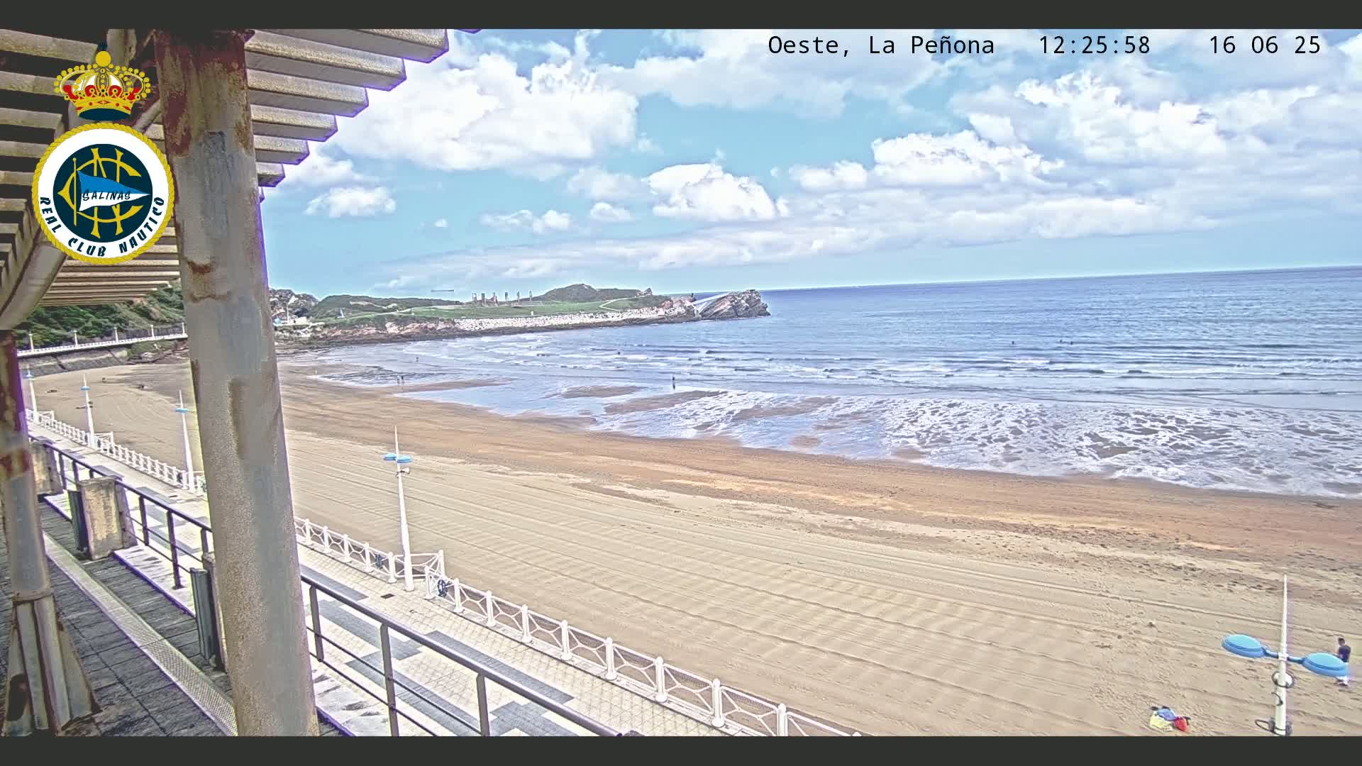 A sandy beach with gently rolling waves under a partly cloudy sky is viewed from an elevated vantage point.