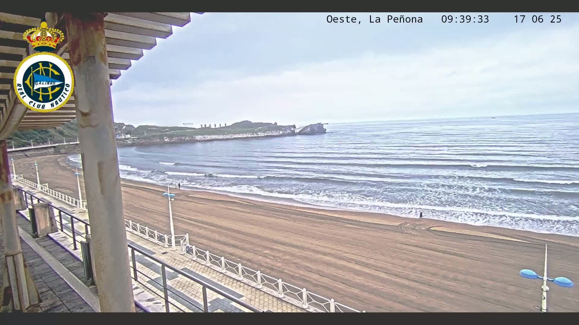 A view from a covered balcony shows a mostly empty sandy beach with gentle waves rolling onto the shore under an overcast sky.
