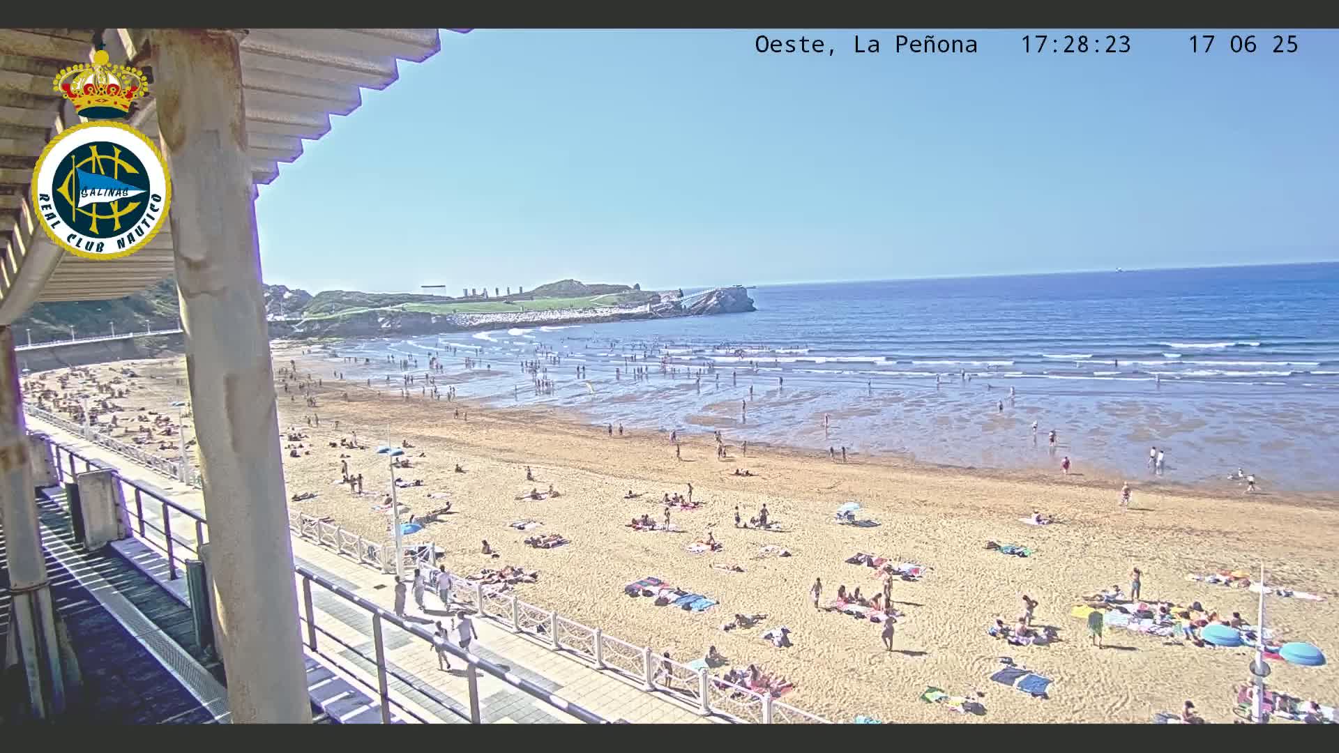 A sandy beach with many people sunbathing and swimming, under a clear blue sky, is seen from an elevated vantage point near a building.