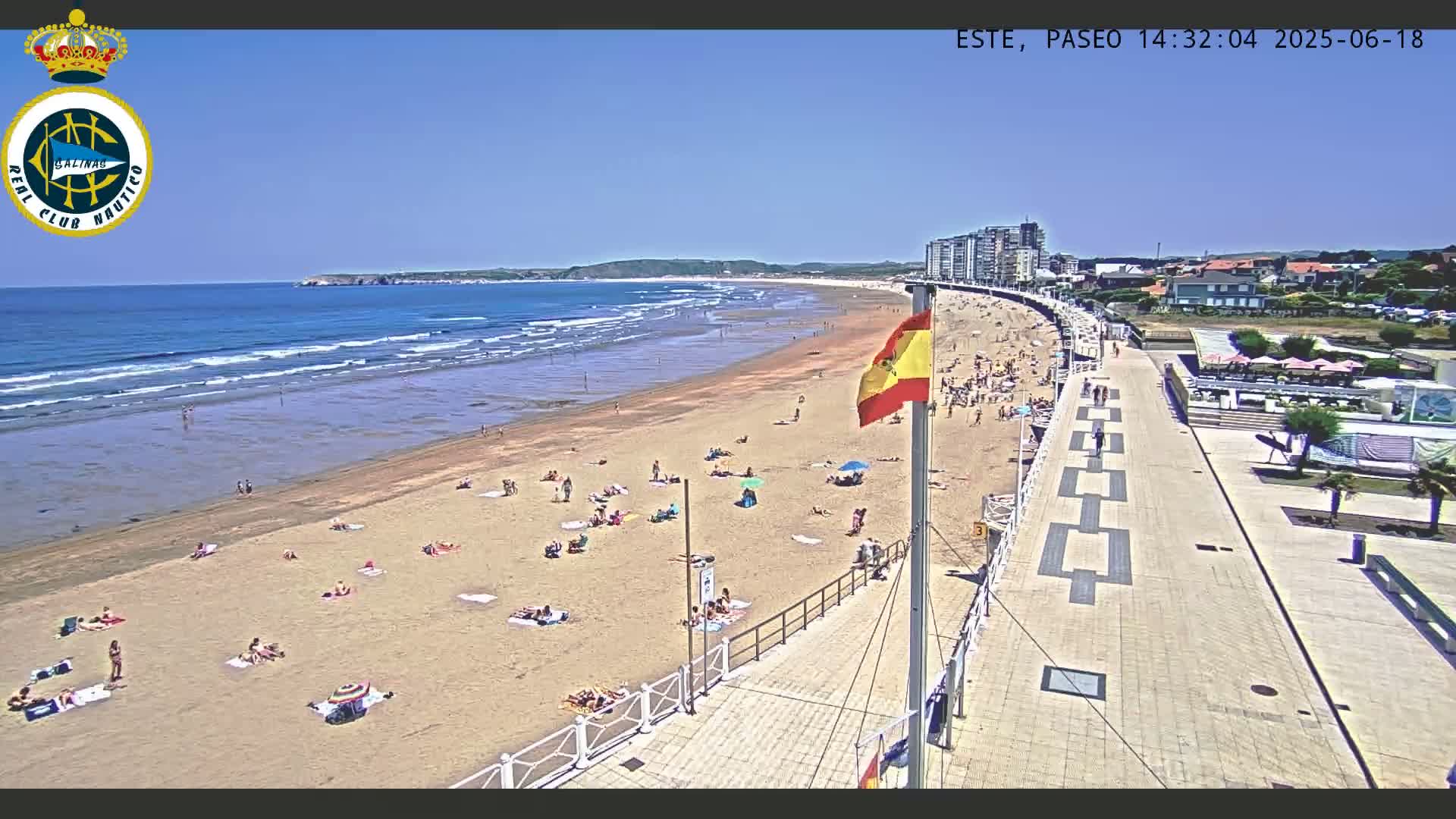 A sandy beach with many people sunbathing and swimming, under a clear blue sky, is bordered by a paved walkway leading to buildings.