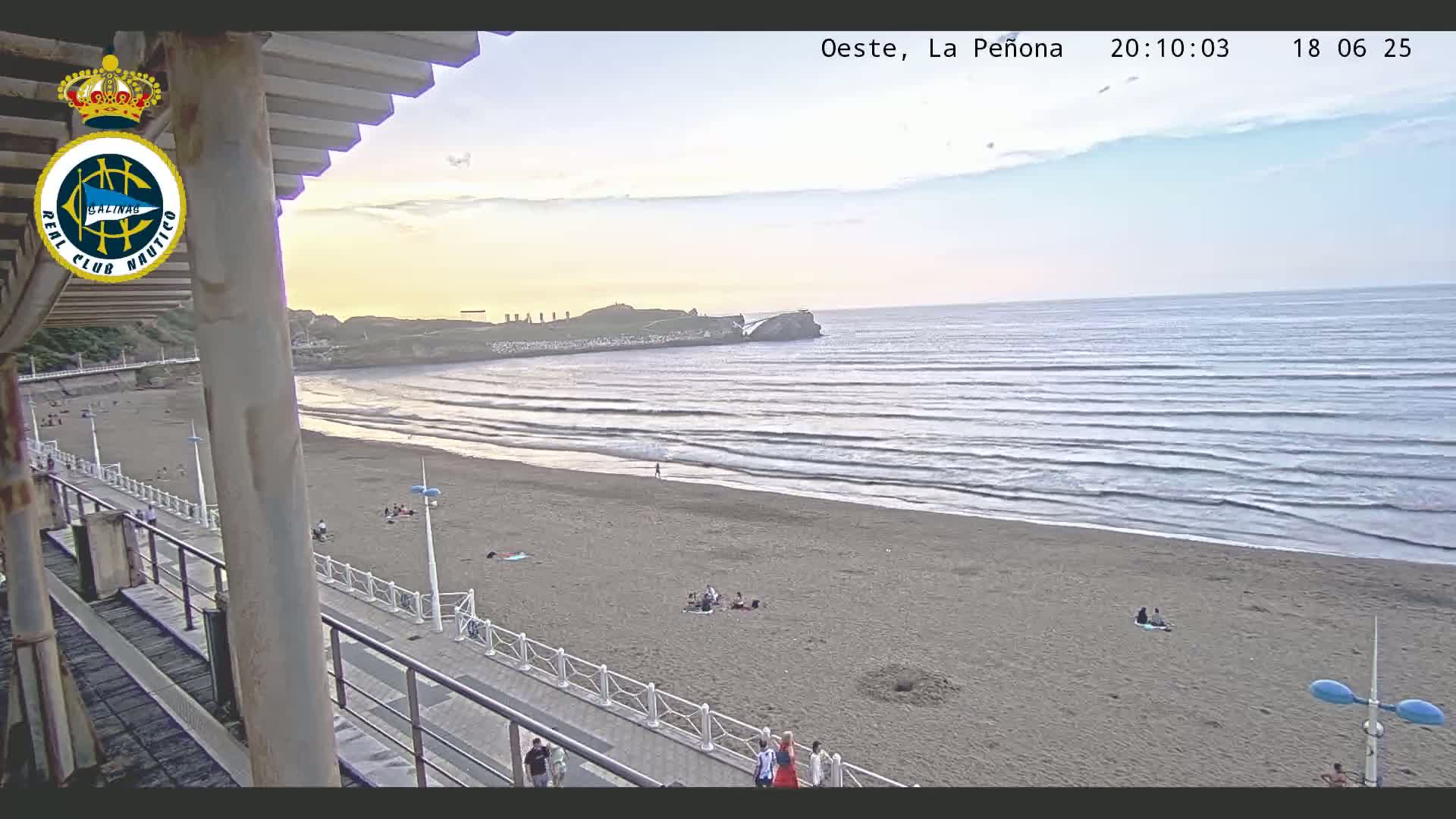 A sandy beach with calm waves and several people are visible at sunset from an elevated vantage point.