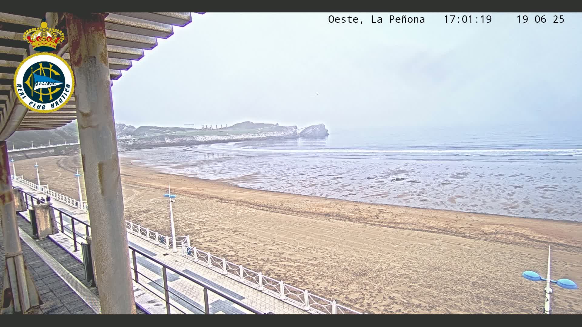 A sandy beach with a calm ocean and a hazy sky is viewed from a boardwalk under a partially visible structure.