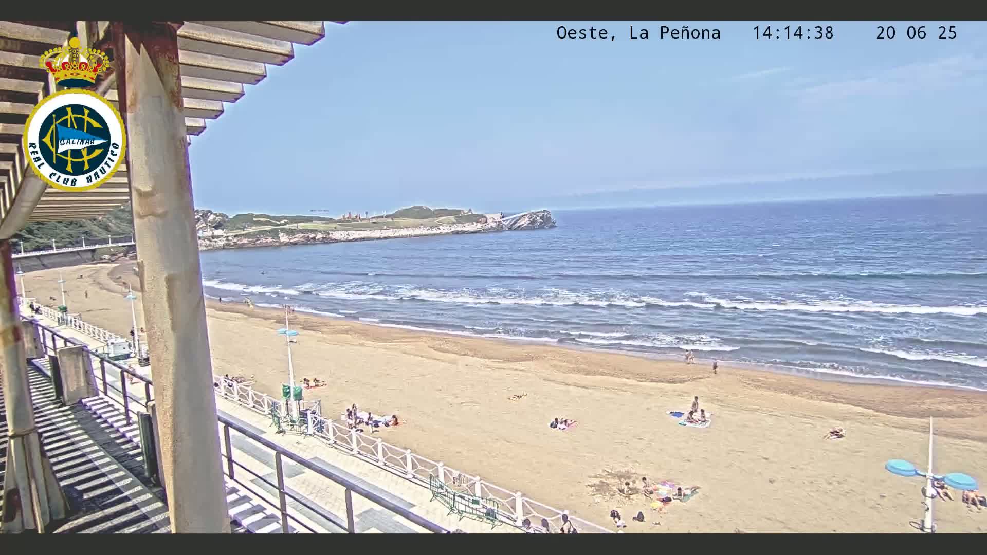 A sandy beach with a few people sunbathing stretches out to a calm ocean under a mostly clear sky, viewed from an elevated vantage point with a walkway and pergola in the foreground.