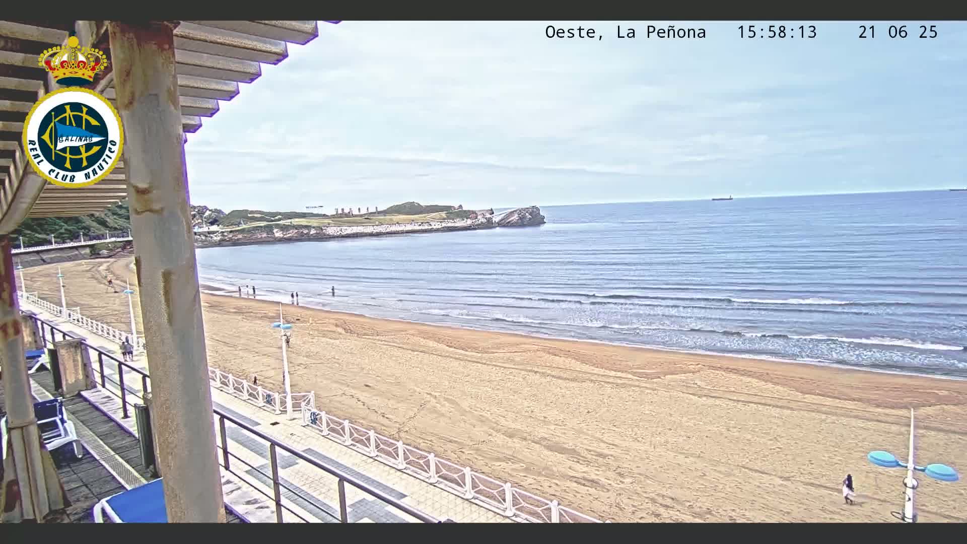 A sandy beach with gentle waves and a few people walking on it stretches out below a partly shaded boardwalk on a partly cloudy day.