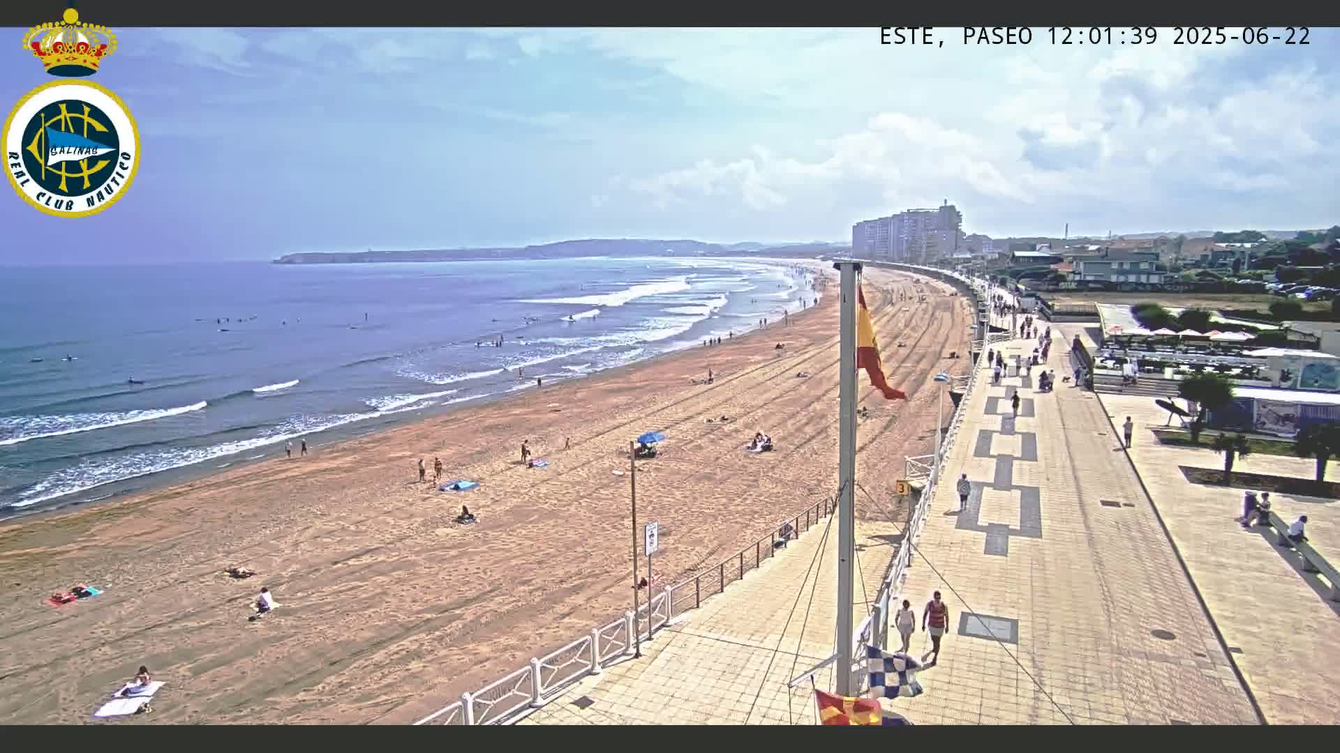 A sandy beach with people sunbathing and swimming in a calm ocean, under a partly cloudy sky, is adjacent to a paved boardwalk with pedestrians.
