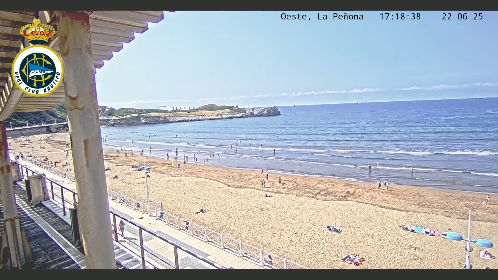 A sandy beach with a moderate number of people, calm ocean waves, and a clear blue sky is viewed from a partially covered elevated vantage point.