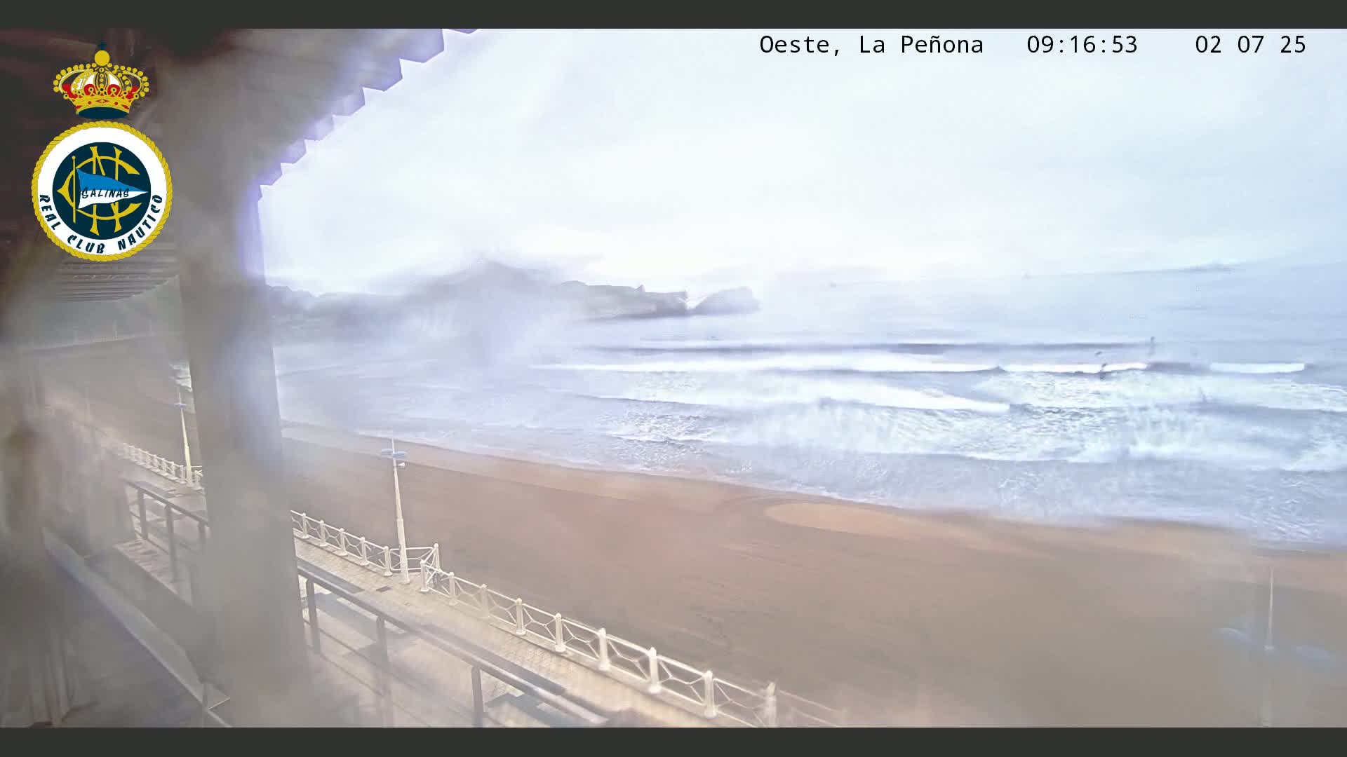 A hazy, overcast day reveals a sandy beach with waves breaking near a shoreline promenade and distant rocky outcrops.