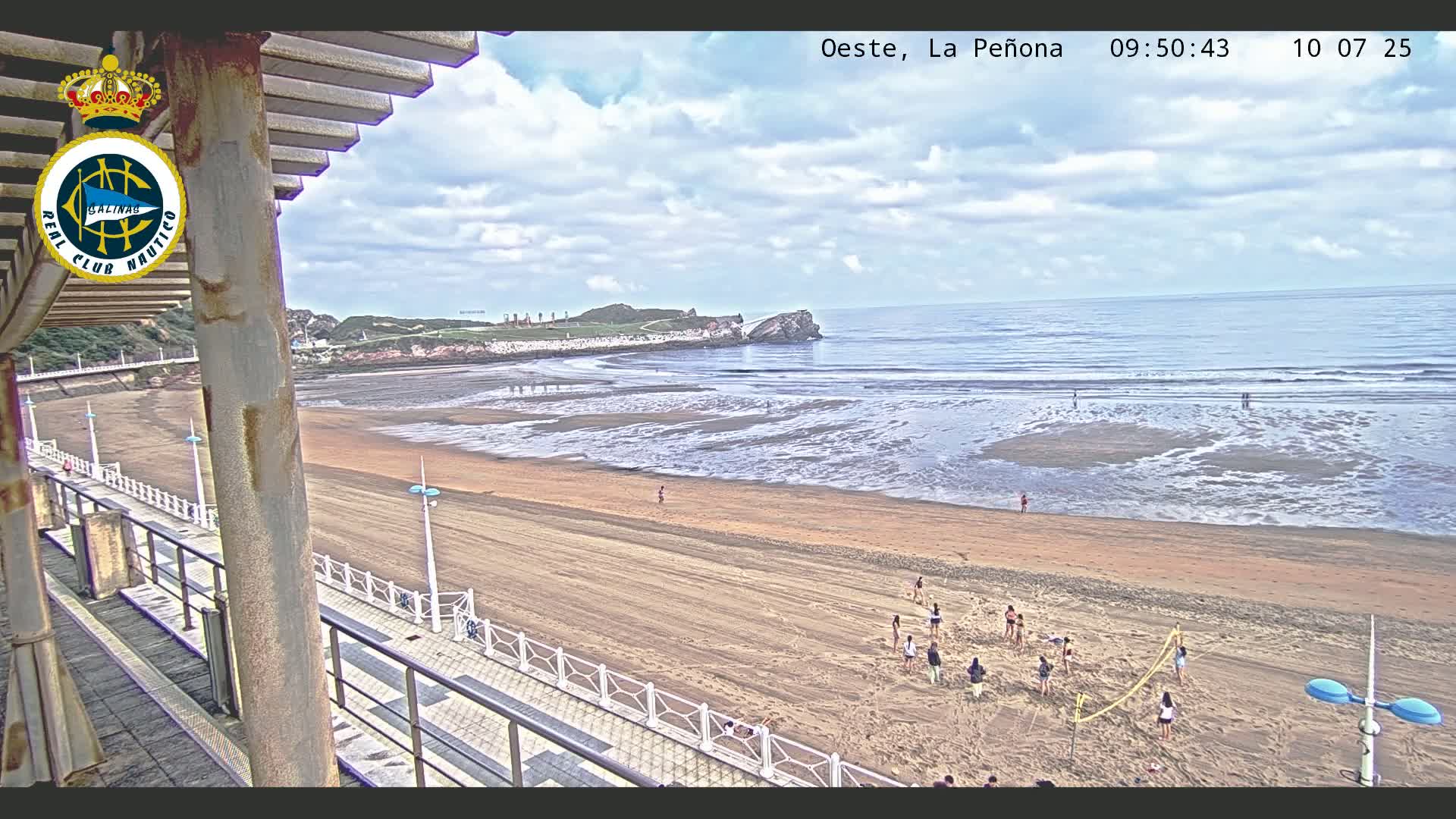 A sandy beach with people playing volleyball stretches out to a calm ocean under a partly cloudy sky.
