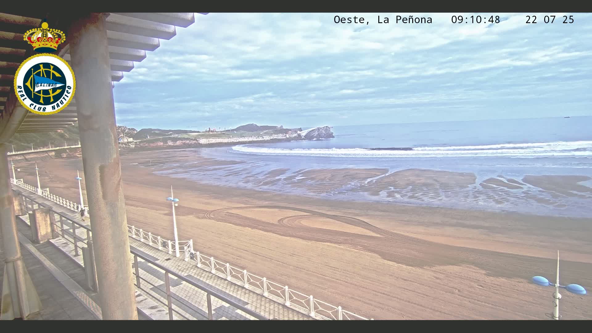 A sandy beach with gentle waves and cloudy skies is viewed from a balcony overlooking a boardwalk.