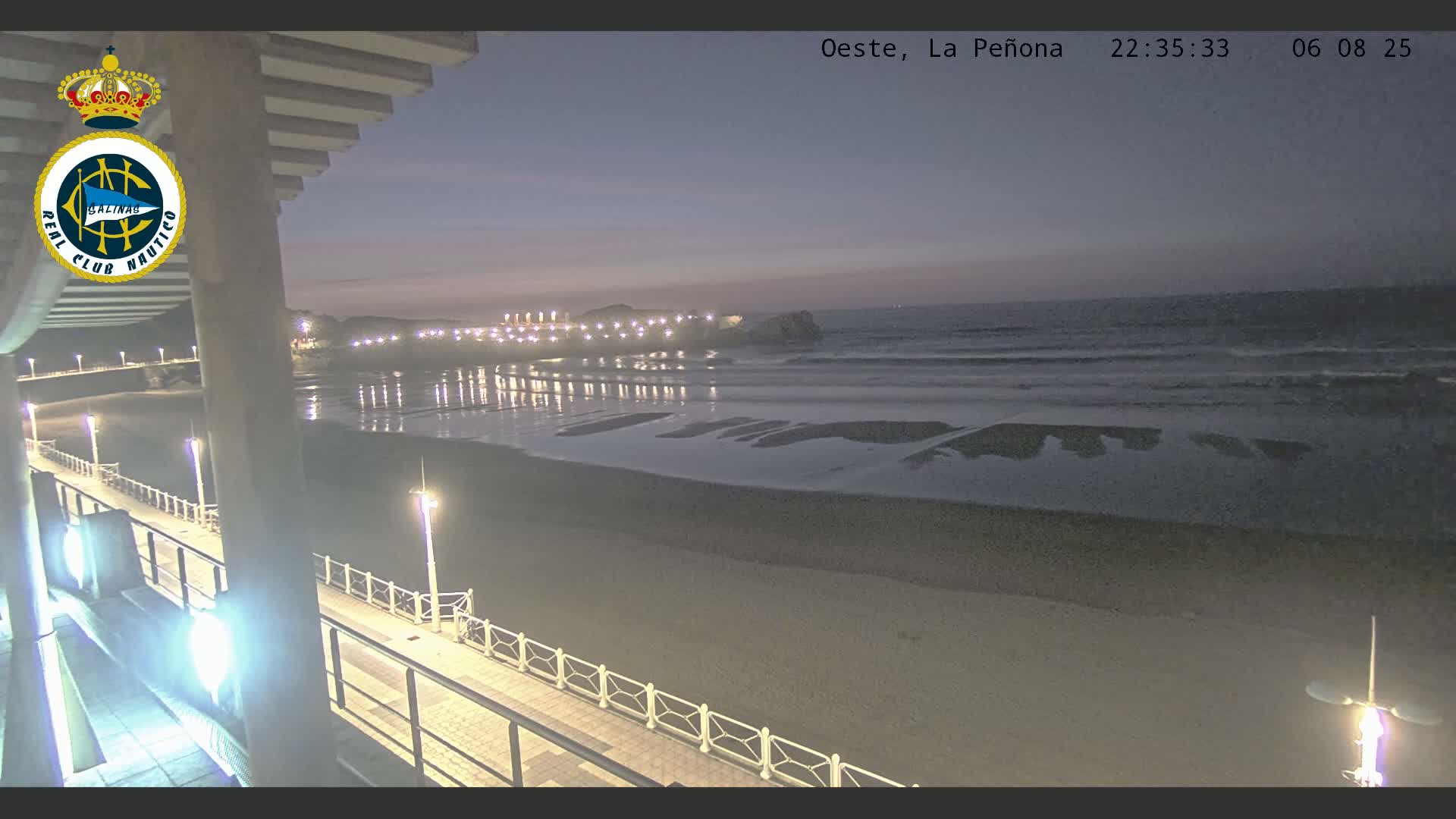 A nighttime view of a beach with gently rolling waves under a dusky sky, showing a pier with lights extending into the water.