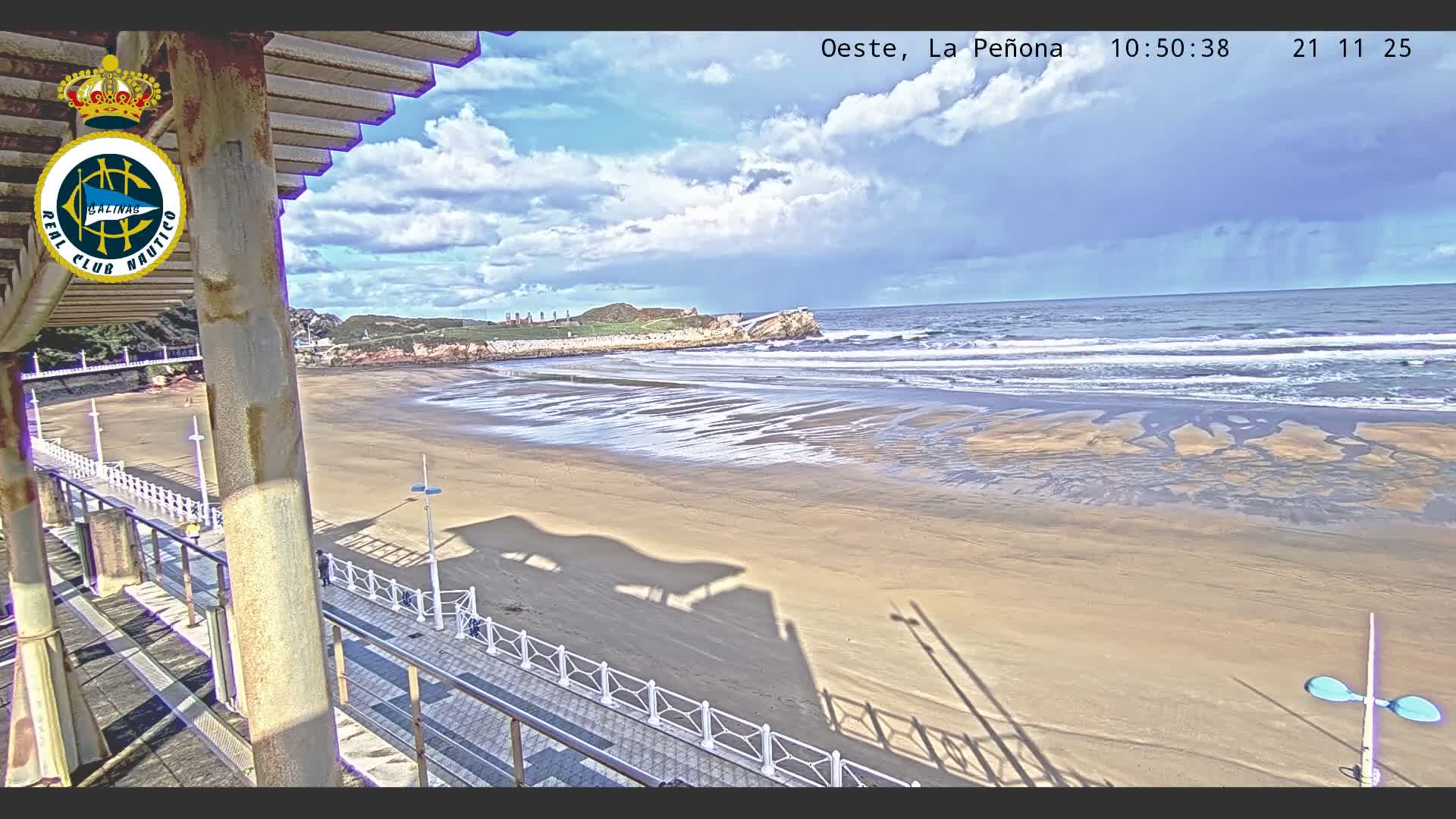 A wide coastal view from a pier reveals a broad sandy beach stretching towards an ocean with breaking waves and a distant rocky headland, all under a partly cloudy sky with prominent shadows.