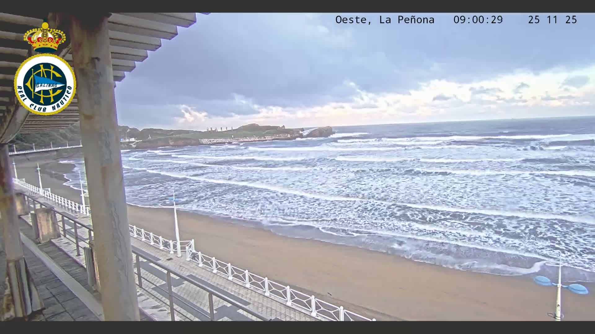 The image displays a wide sandy beach with moderate waves breaking under a heavily overcast sky, viewed from an elevated perspective next to a pier with white railings, looking towards a rocky coastline in the distance.