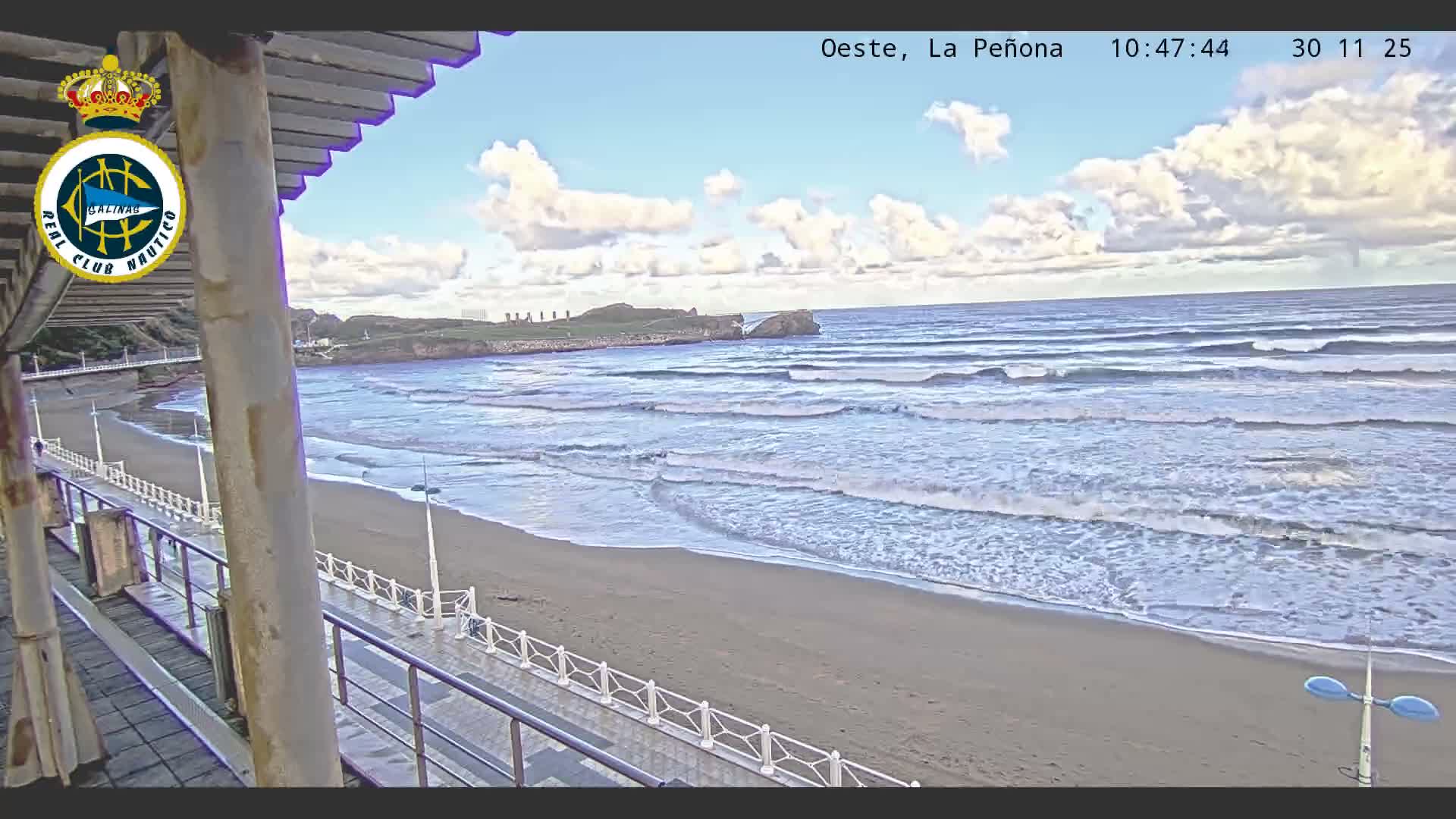 A panoramic view reveals a partly cloudy sky over a wide sandy beach with numerous waves rolling in from the ocean, bordered by a promenade with railings and a distant rocky coastline.