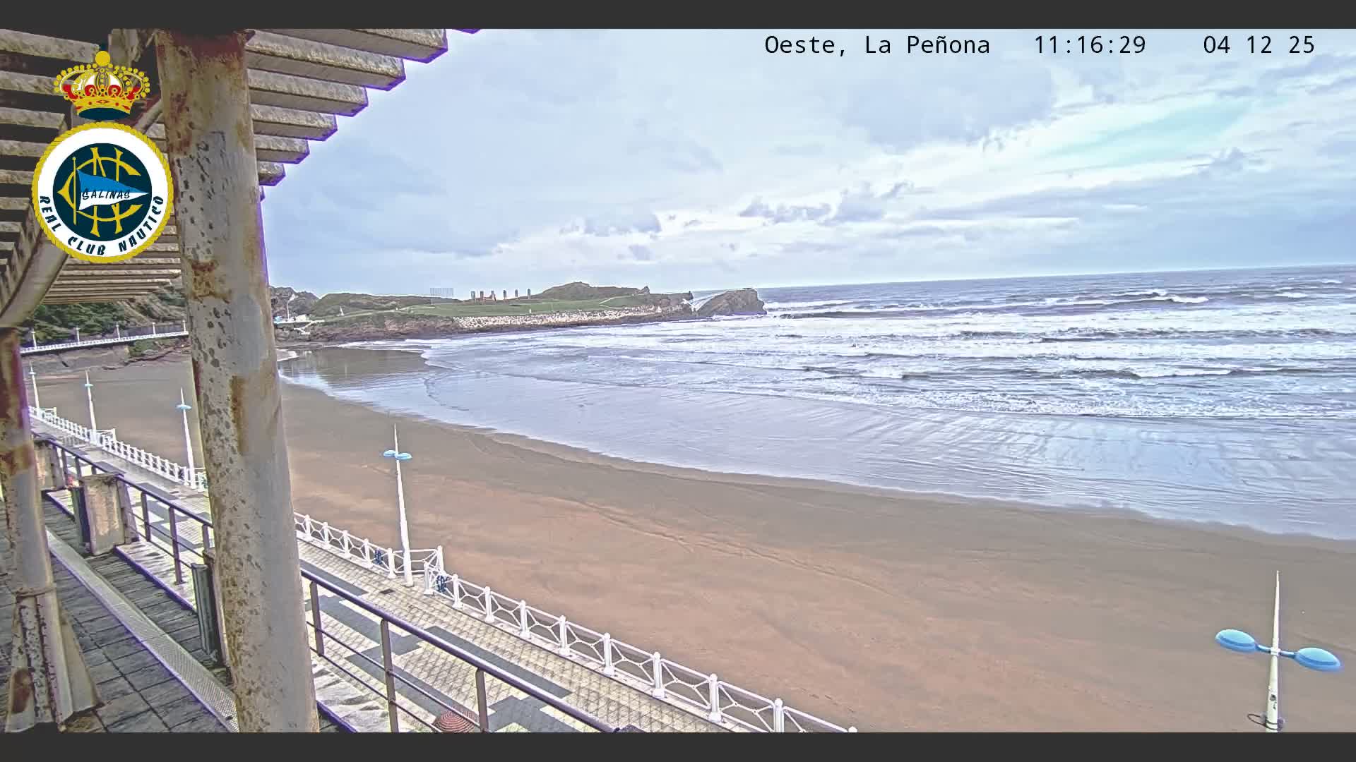 An overcast sky hangs over a wide, sandy beach with breaking waves, viewed from an elevated promenade featuring railings and rusty supports in the foreground, with a rocky headland visible in the distance.