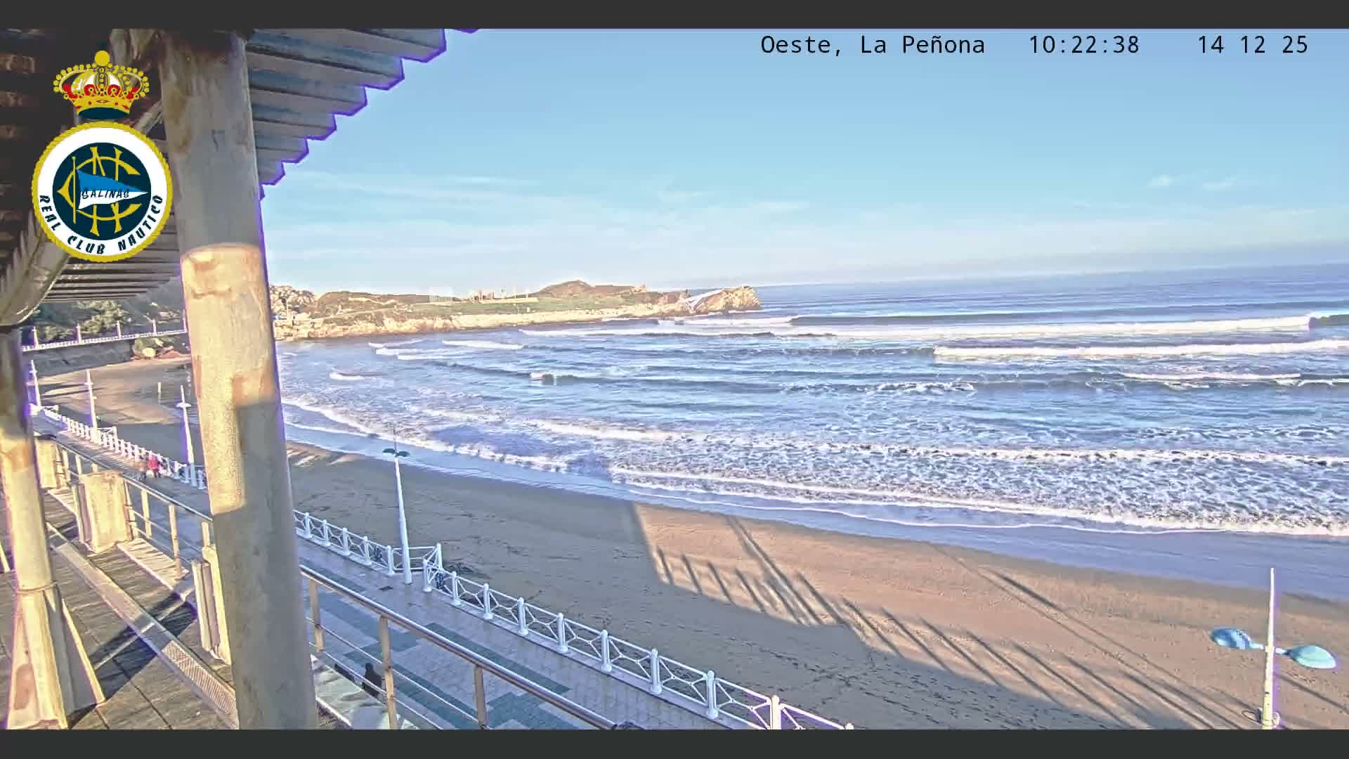 An overcast sky hangs over a wide, sandy beach with breaking waves, viewed from an elevated promenade featuring railings and rusty supports in the foreground, with a rocky headland visible in the distance.
