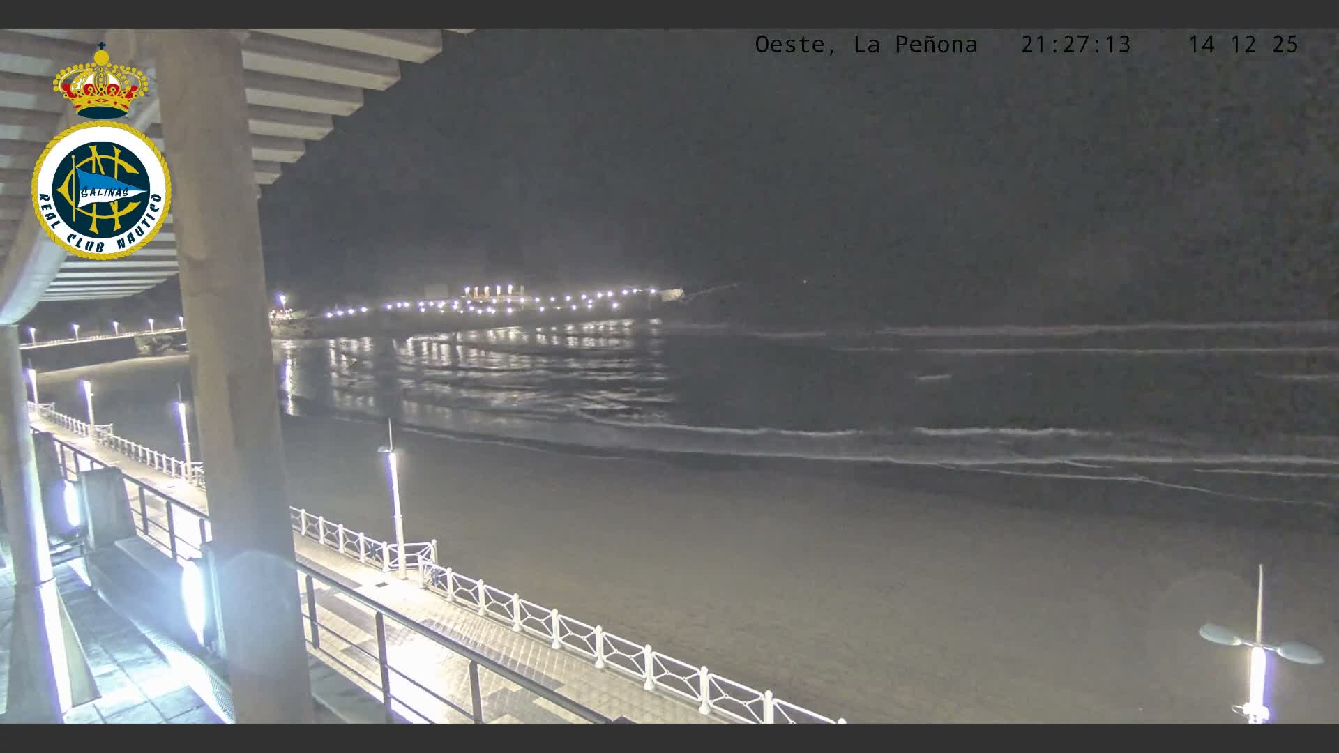 An overcast sky hangs over a wide, sandy beach with breaking waves, viewed from an elevated promenade featuring railings and rusty supports in the foreground, with a rocky headland visible in the distance.