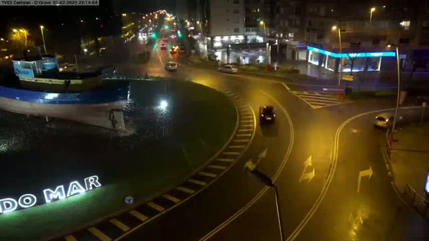 A wet roundabout at night with a few cars and a model of a boat in the center.